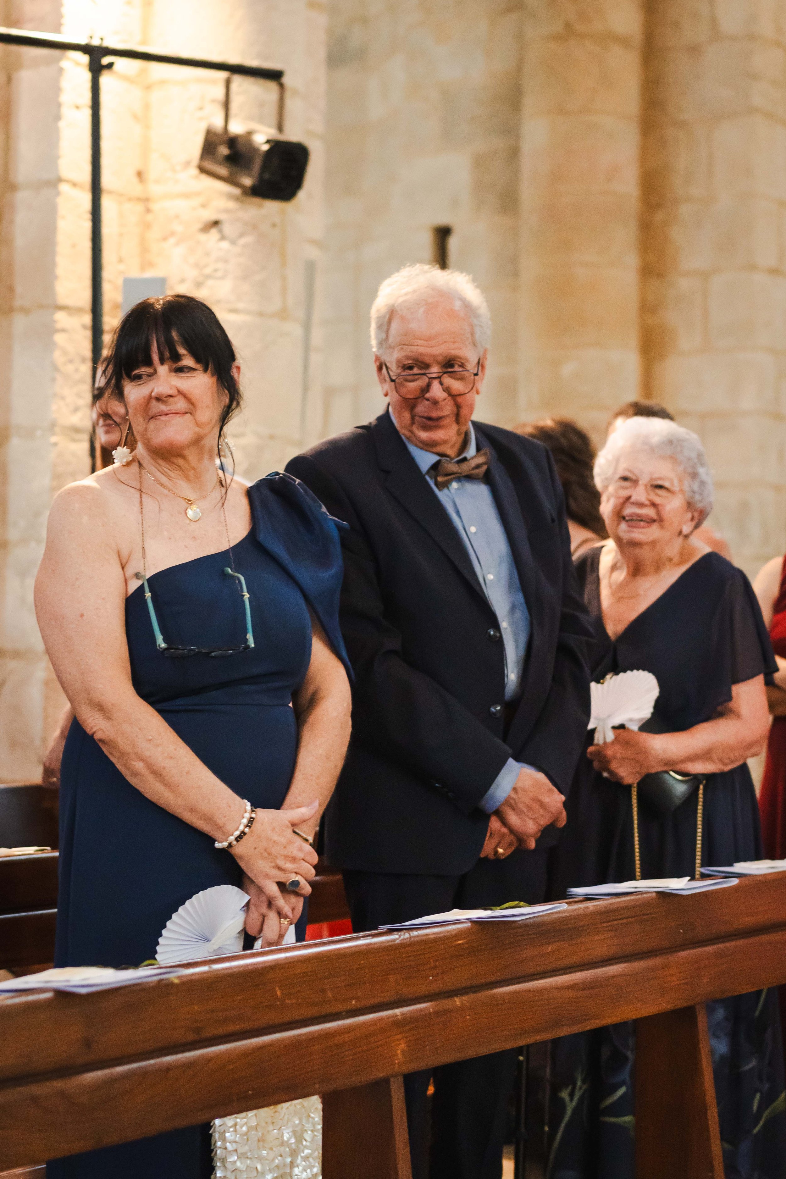 Trois personnes âgées en tenue formelle dans une église, probablement lors d'une cérémonie ou d'un mariage.