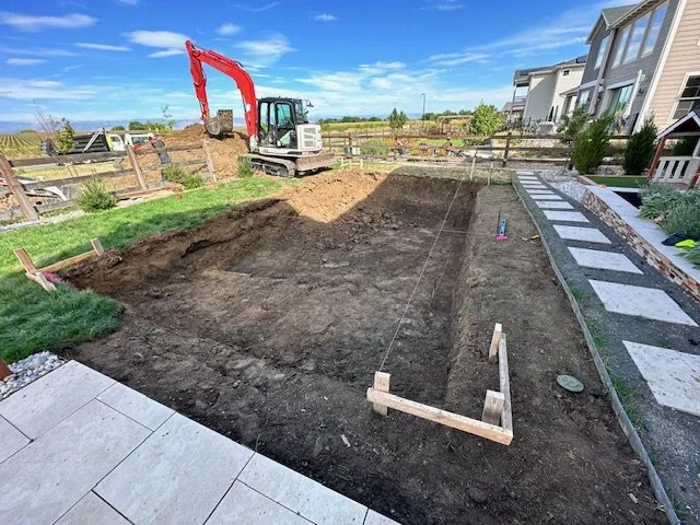 A backyard in a residential area with a large empty garden bed, construction equipment, and a row of townhouses on the right.