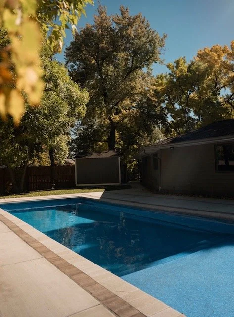 Residential backyard with a swimming pool, surrounded by trees and a house in the background.