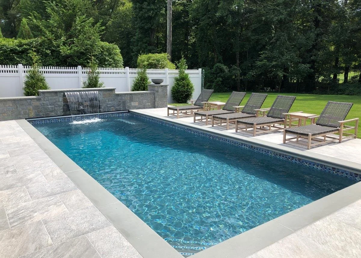 A backyard swimming pool with a waterfall feature, surrounded by a light-colored tiled deck. Several lounge chairs with mesh seats are lined up along the poolside, and there is a white picket fence with bushes behind it. In the background, there are trees and greenery.
