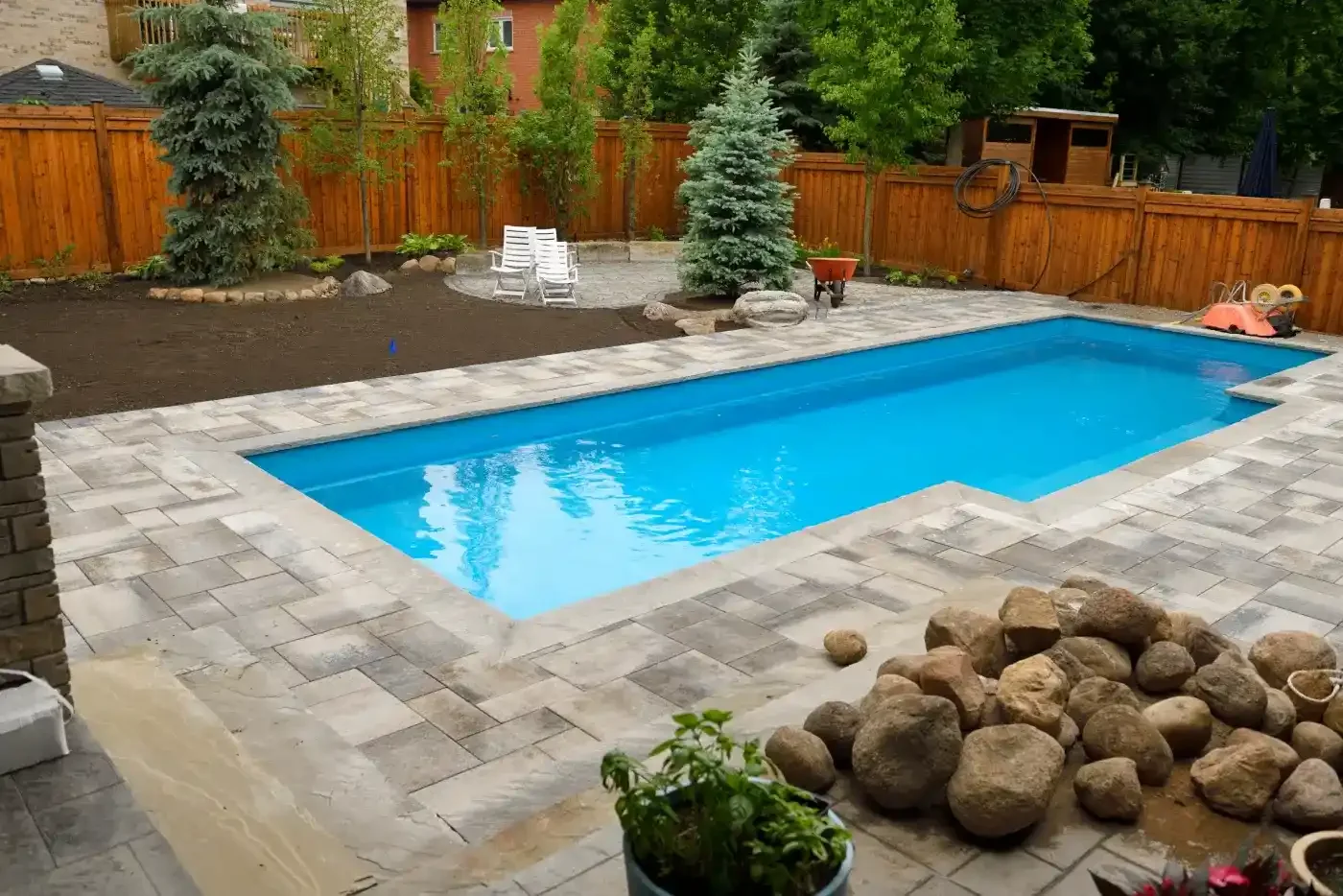 Backyard with a swimming pool, stone patio, trees, and a wooden fence.