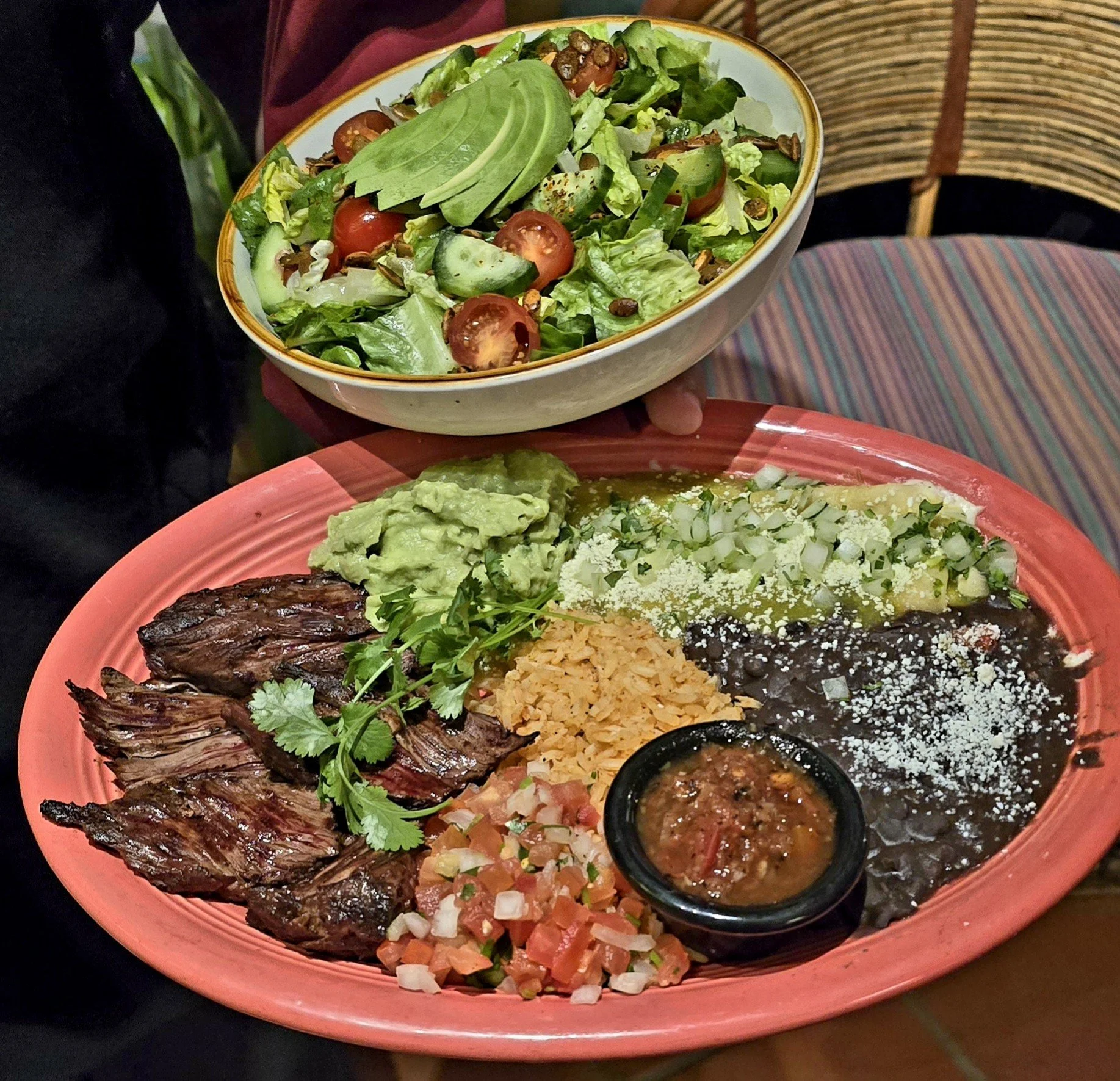 A plate of Mexican food with grilled meat, guacamole, rice, black beans with cheese, chopped onions and tomatoes, and green sauce, with a salad of lettuce, cherry tomatoes, cucumbers, avocado slices, and pumpkin seeds