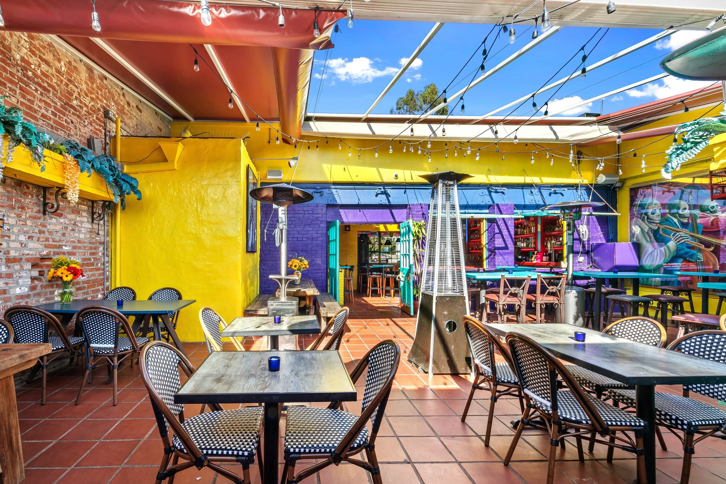 Colorful outdoor patio with yellow, purple, and turquoise walls, string lights, potted flowers, tables and chairs, patio heaters, and a mural of skeleton musicians playing instruments.
