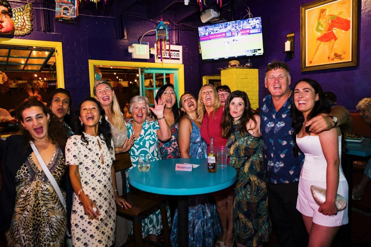 Group of people celebrating at a lively party or bar, smiling and posing for a photo around a table with drinks, colorful decor, and a television screen in the background.