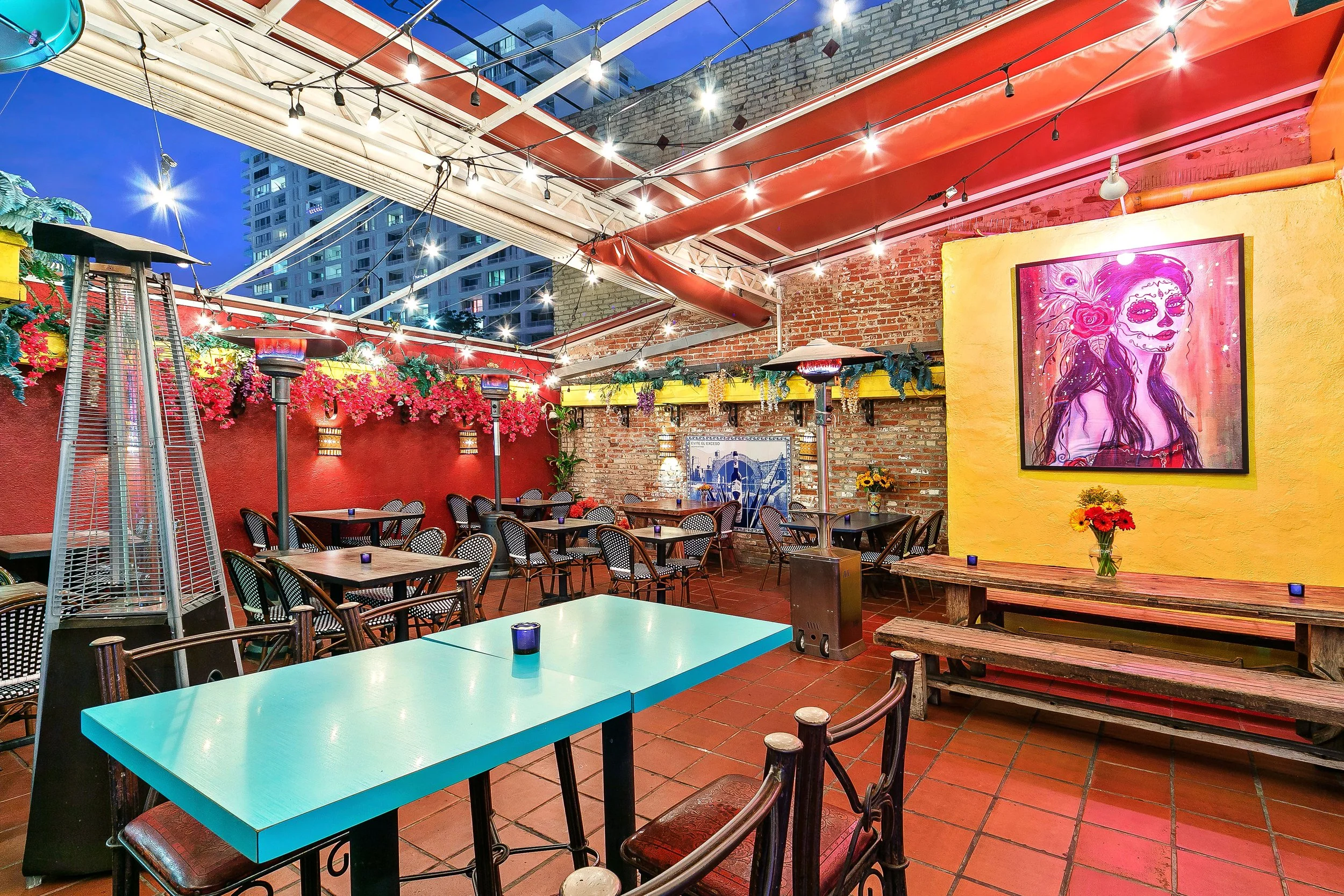 Colorful outdoor patio with string lights, tables, and chairs, decorated wall with flowers and a large colorful artwork of a woman with sugar skull face paint.
