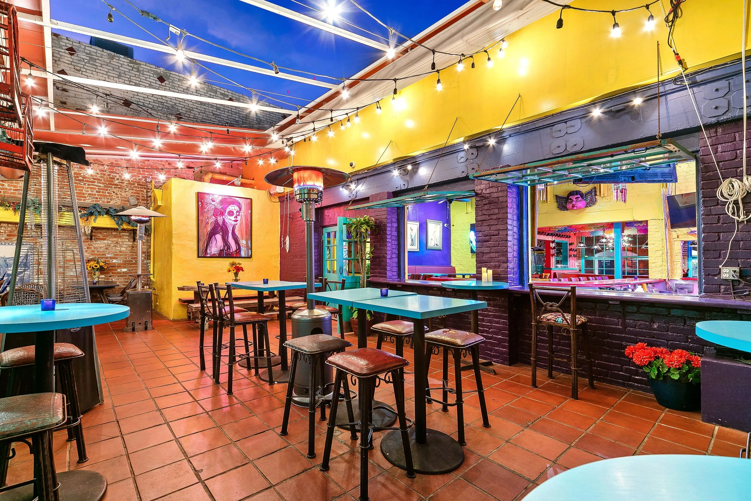Colorful Mexican restaurant with string lights, painted walls, and artwork including a woman painted with sugar skull makeup, brick and painted walls, tables, and chairs.