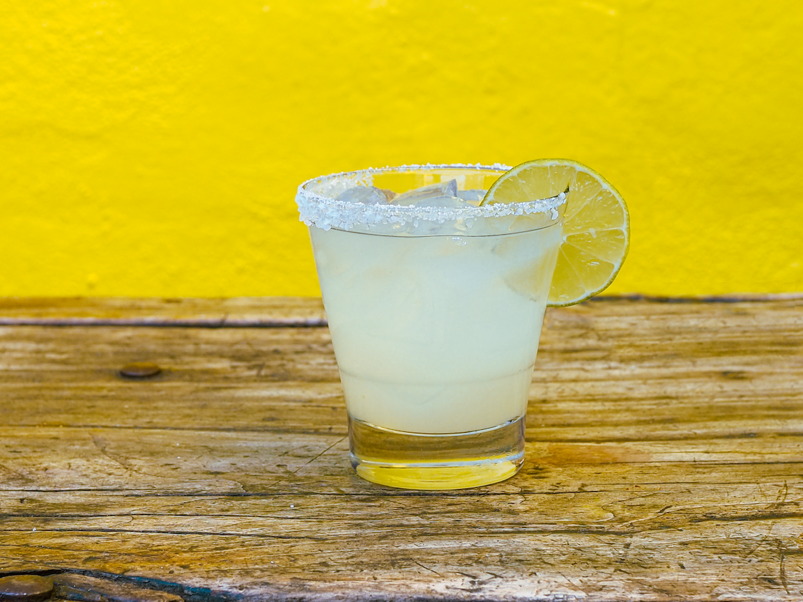 Lula's signature margarita with ice cubes and a lemon slice on the rim, on a rustic wooden table against a yellow background.