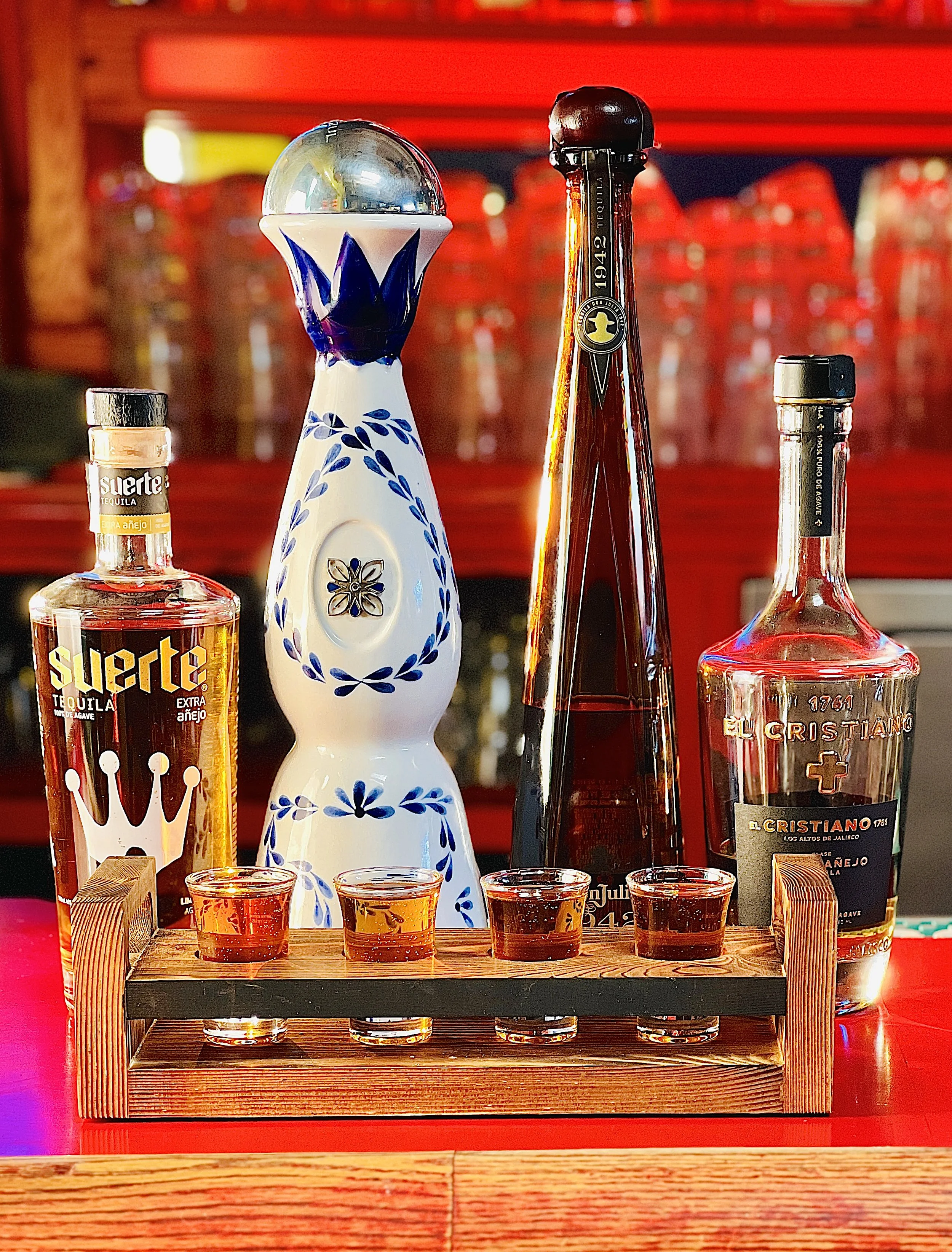 Four tequila bottles and four shot glasses filled with tequila on a wooden tray, with a decorative ceramic pitcher in the background.