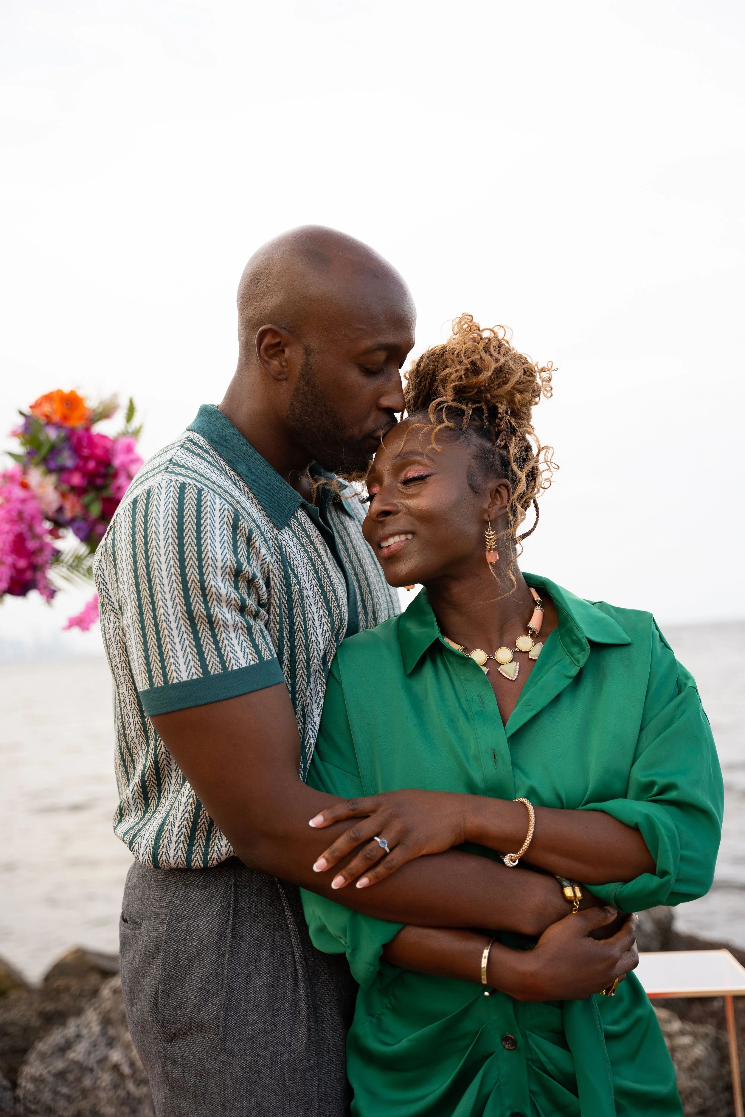 A couple standing close together outdoors by the water, embracing affectionately, with a background of colorful flowers and a cloudy sky.