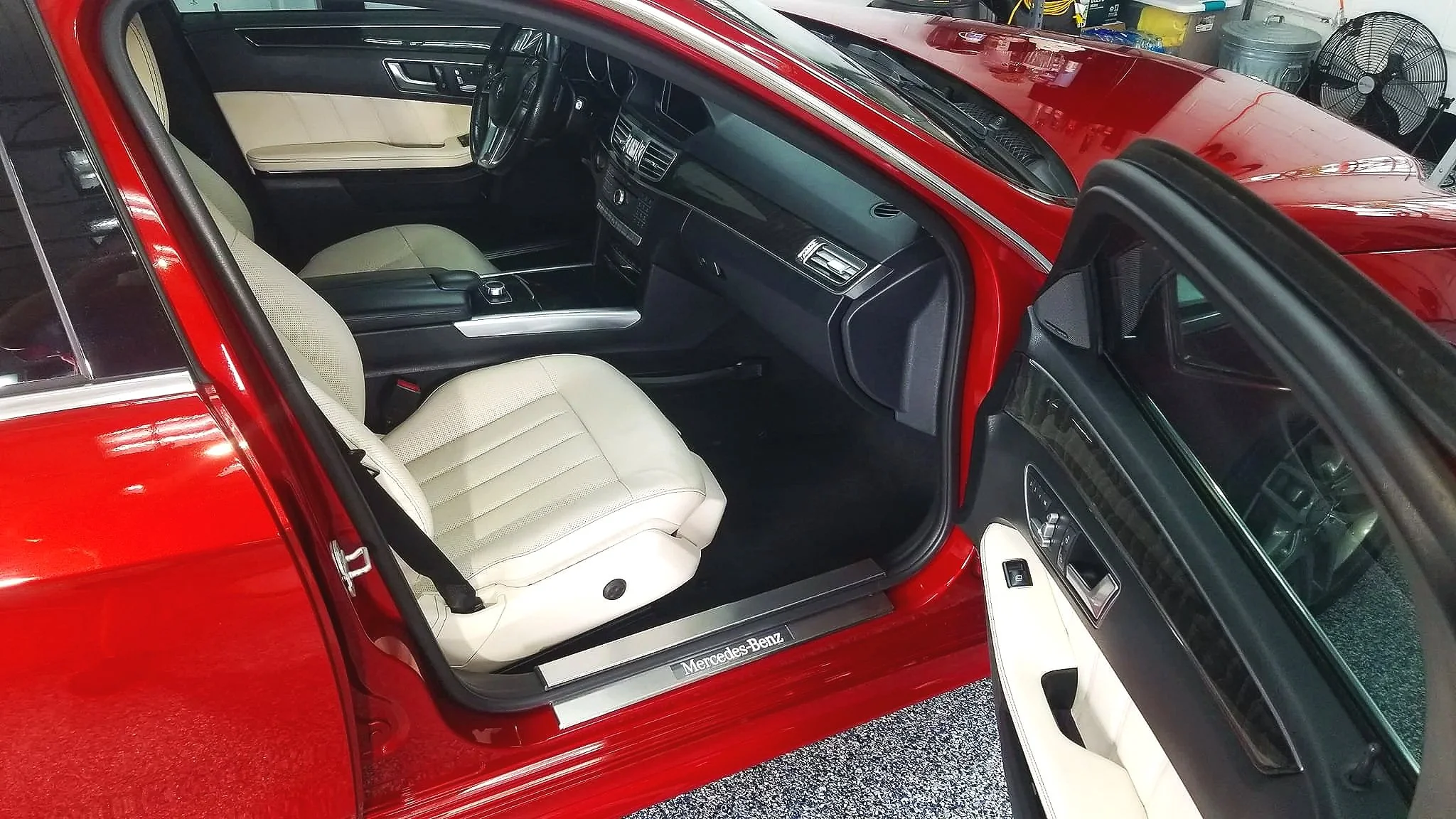Interior of a red Mercedes-Benz car showing a cream-colored seat and a dashboard with black accents, parked in a garage with various household items and a fan in the background.