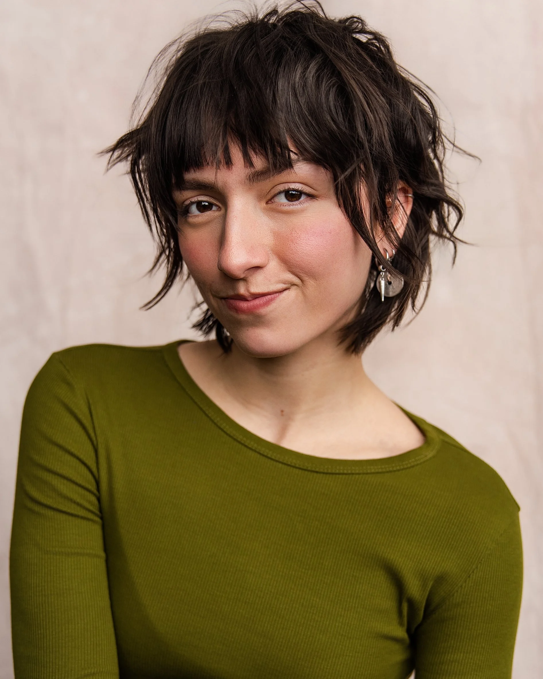 A woman with short, wavy brown hair, wearing a green top and earrings, smiles slightly for the camera against a neutral background.