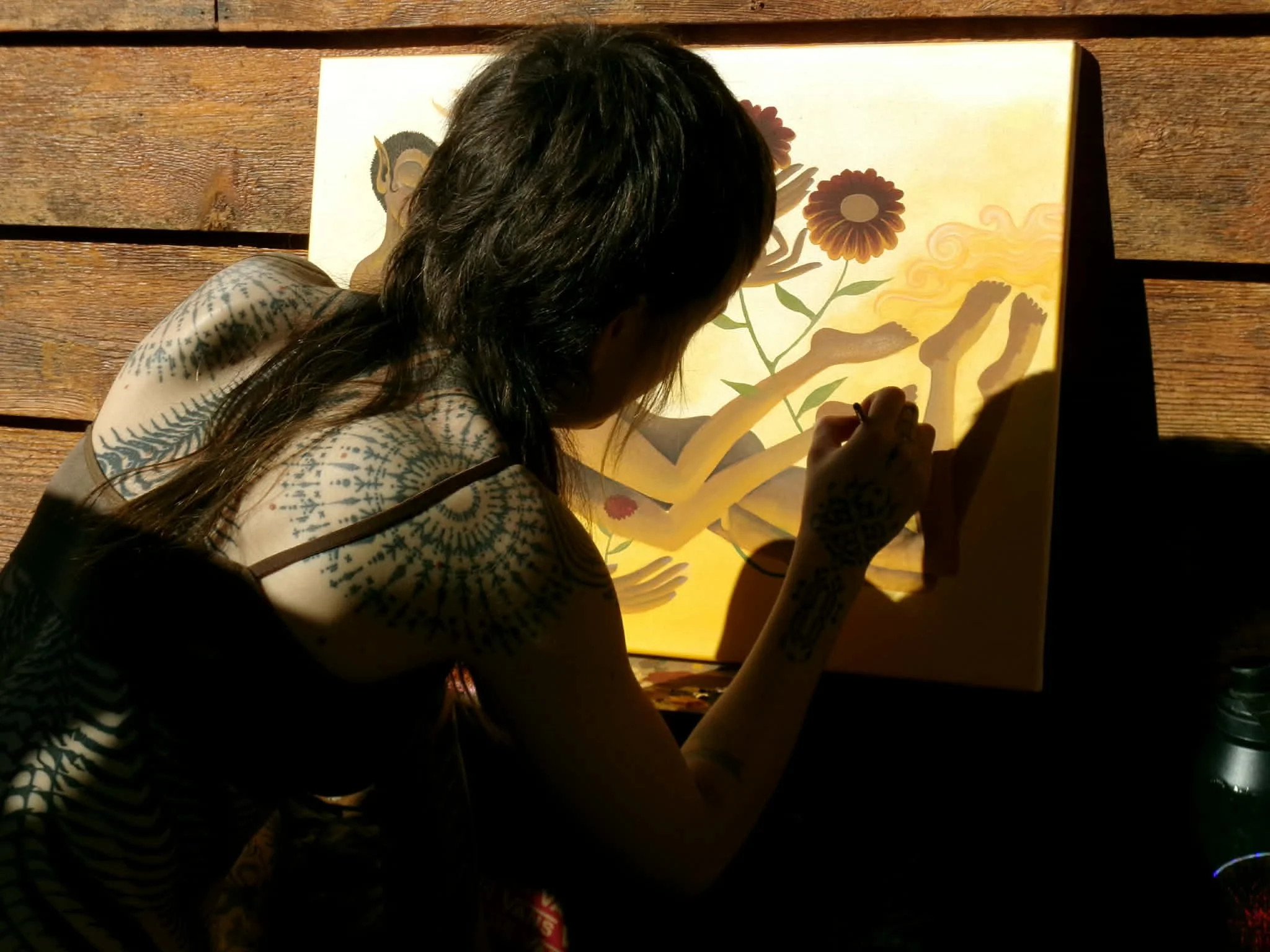 A person with long dark hair, tattoos on their arms, wearing a patterned shirt, sitting at a table and drawing or coloring a picture of a skeleton holding two flowers on a backlit lightbox.