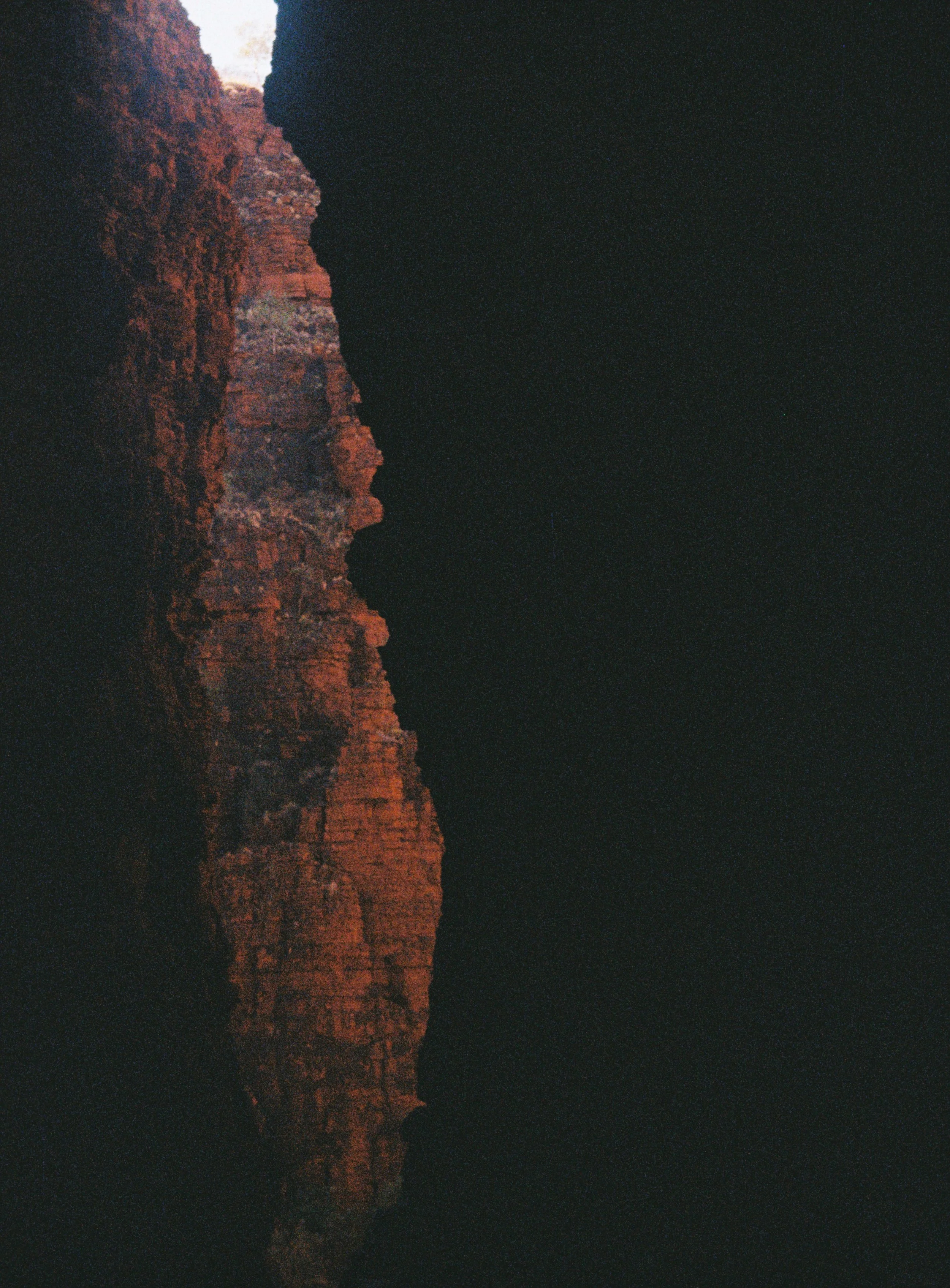 Nighttime view of a red rock cliff illuminated with a faint glow, reflected in a lake with a dark sky in the background.