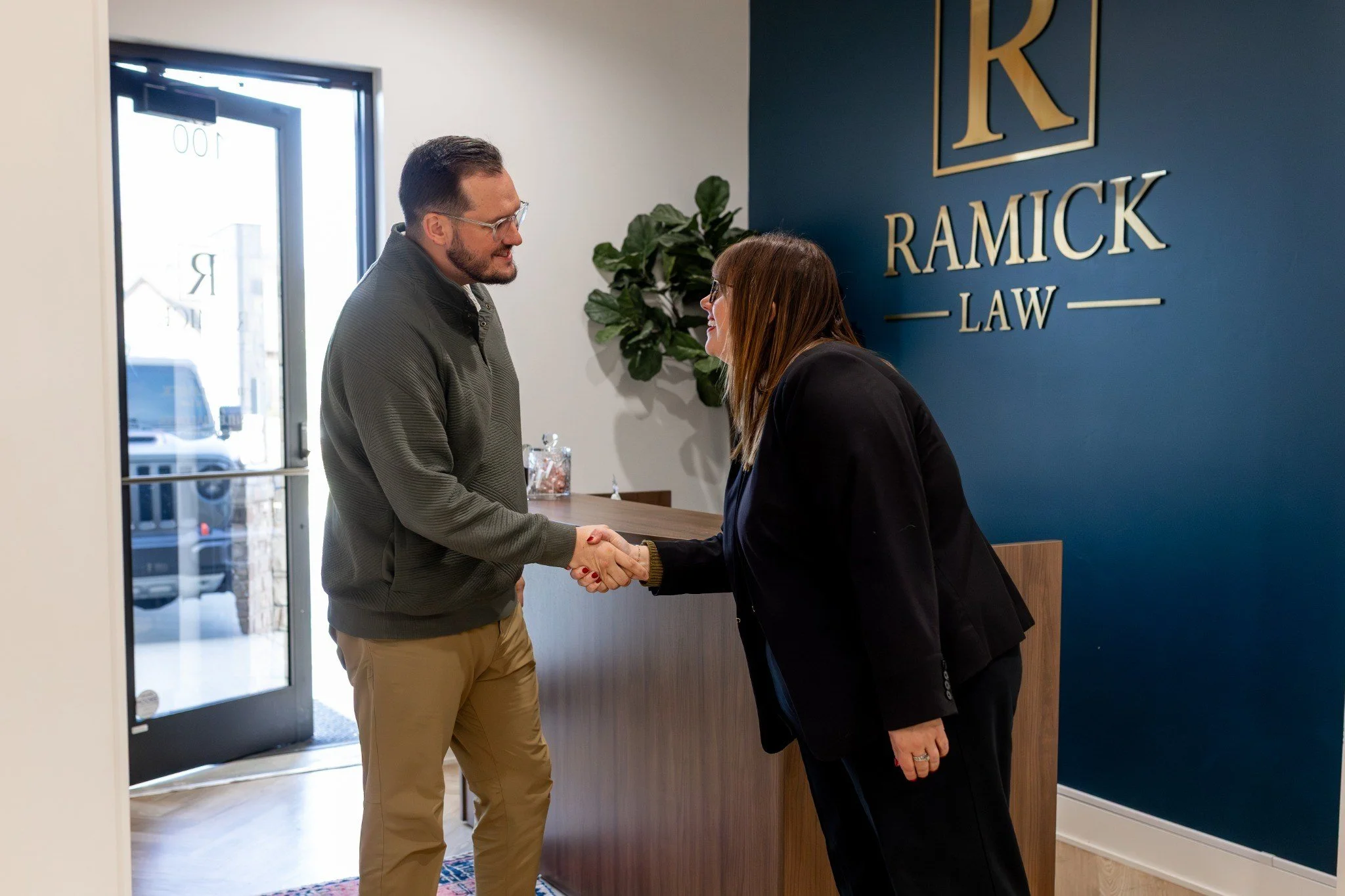 A man and woman shake hands in a law office reception area. The man wears glasses, a gray jacket, and khakis. The woman has long brown hair, glasses, and a black blazer. Behind them is a blue wall with a large gold sign reading 'RAMICK LAW' and a potted plant nearby.