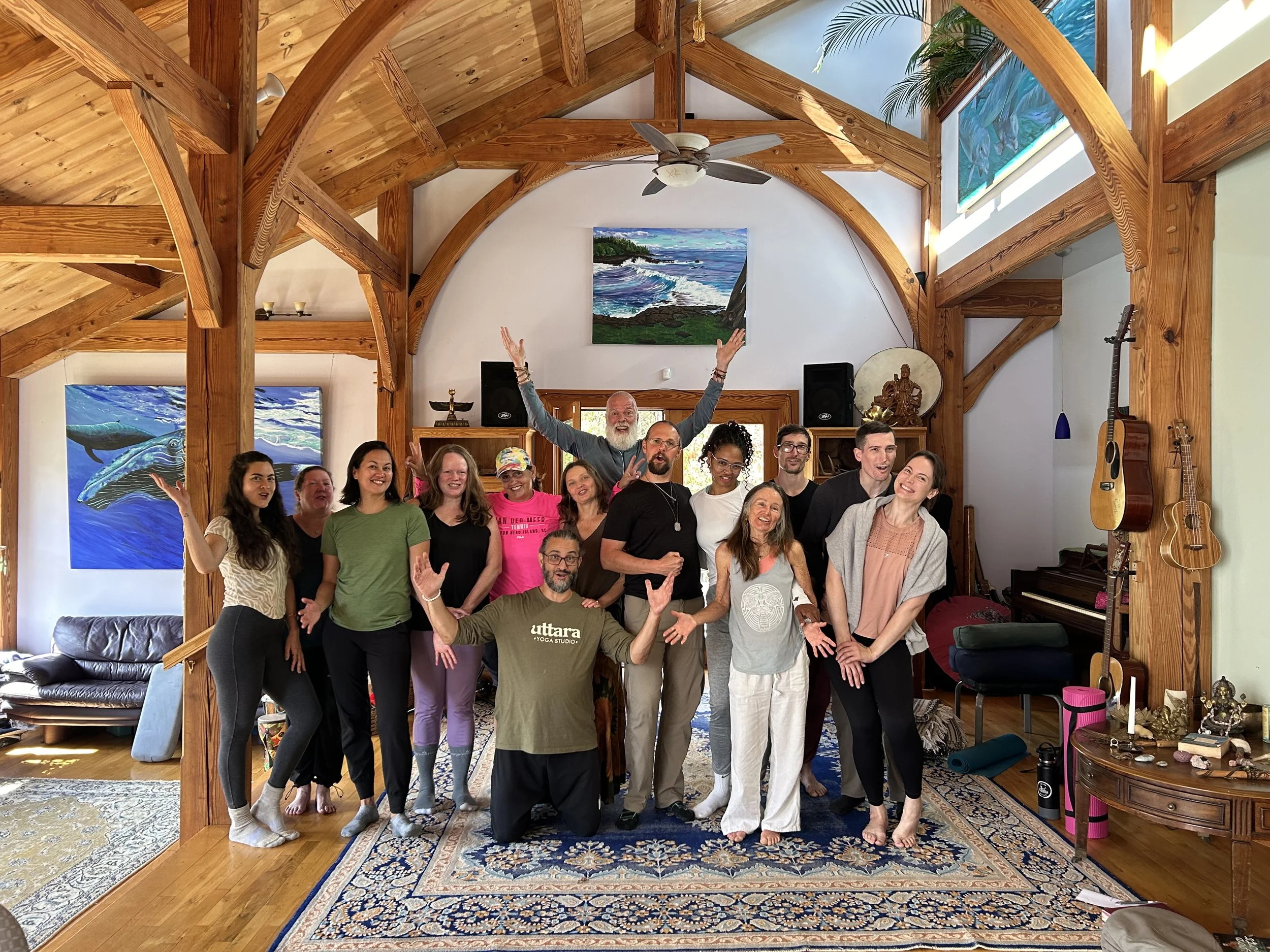 A group of people standing together inside a room with wooden beams, colorful artwork on the walls, and musical instruments like guitars and a piano. They are smiling and making gestures of celebration.
