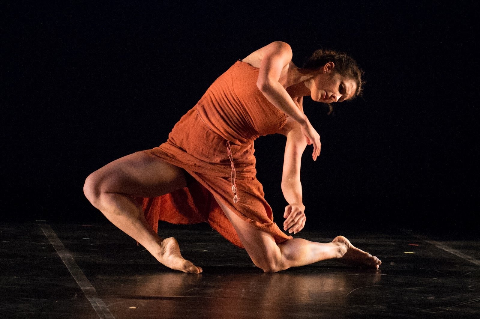 A female dancer in a burnt orange dress performing a contemporary dance move on a dark stage.