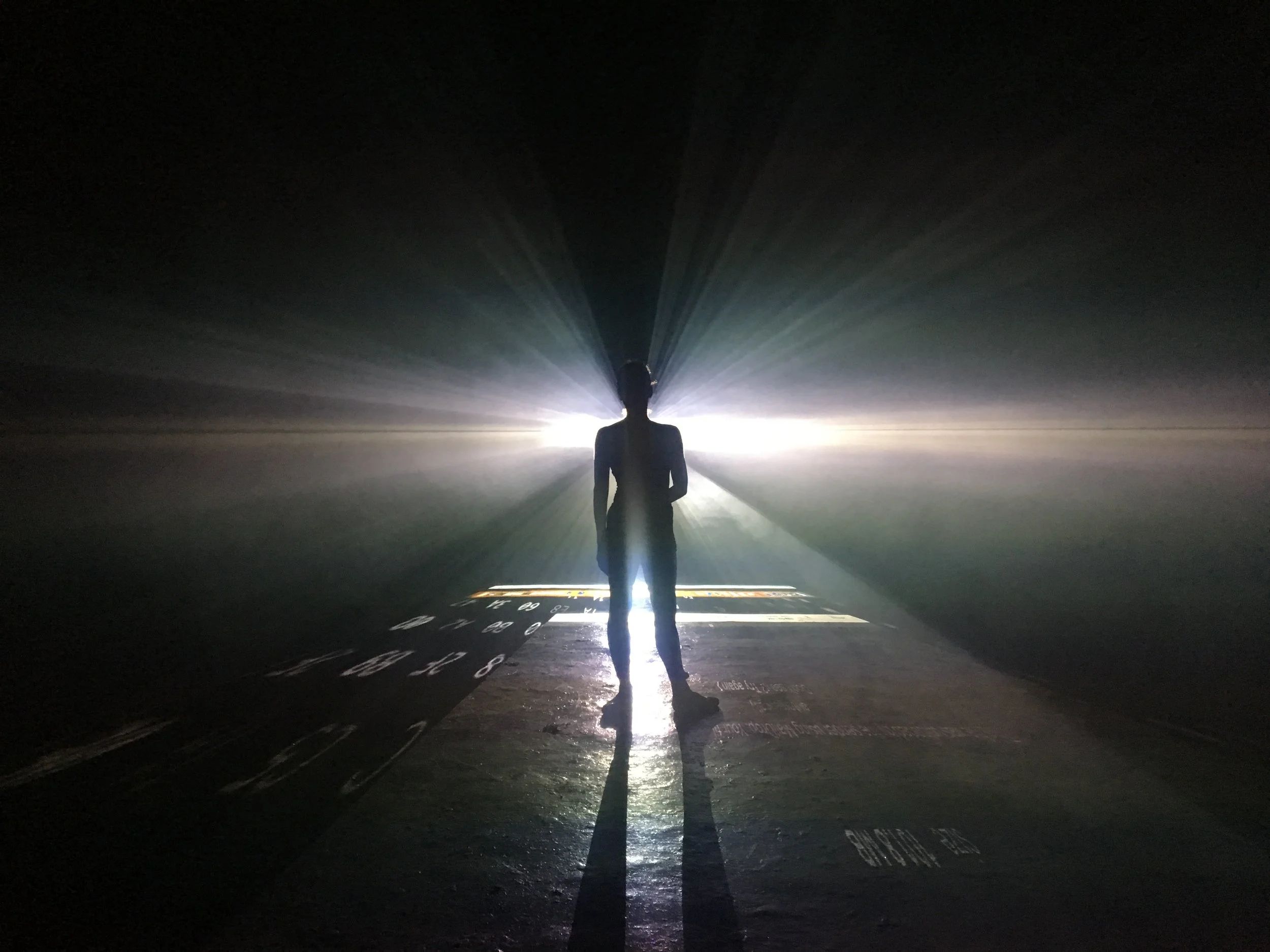 A person standing in a dark room illuminated from behind by bright light, with rays emanating outward and their shadow cast forward on the floor.