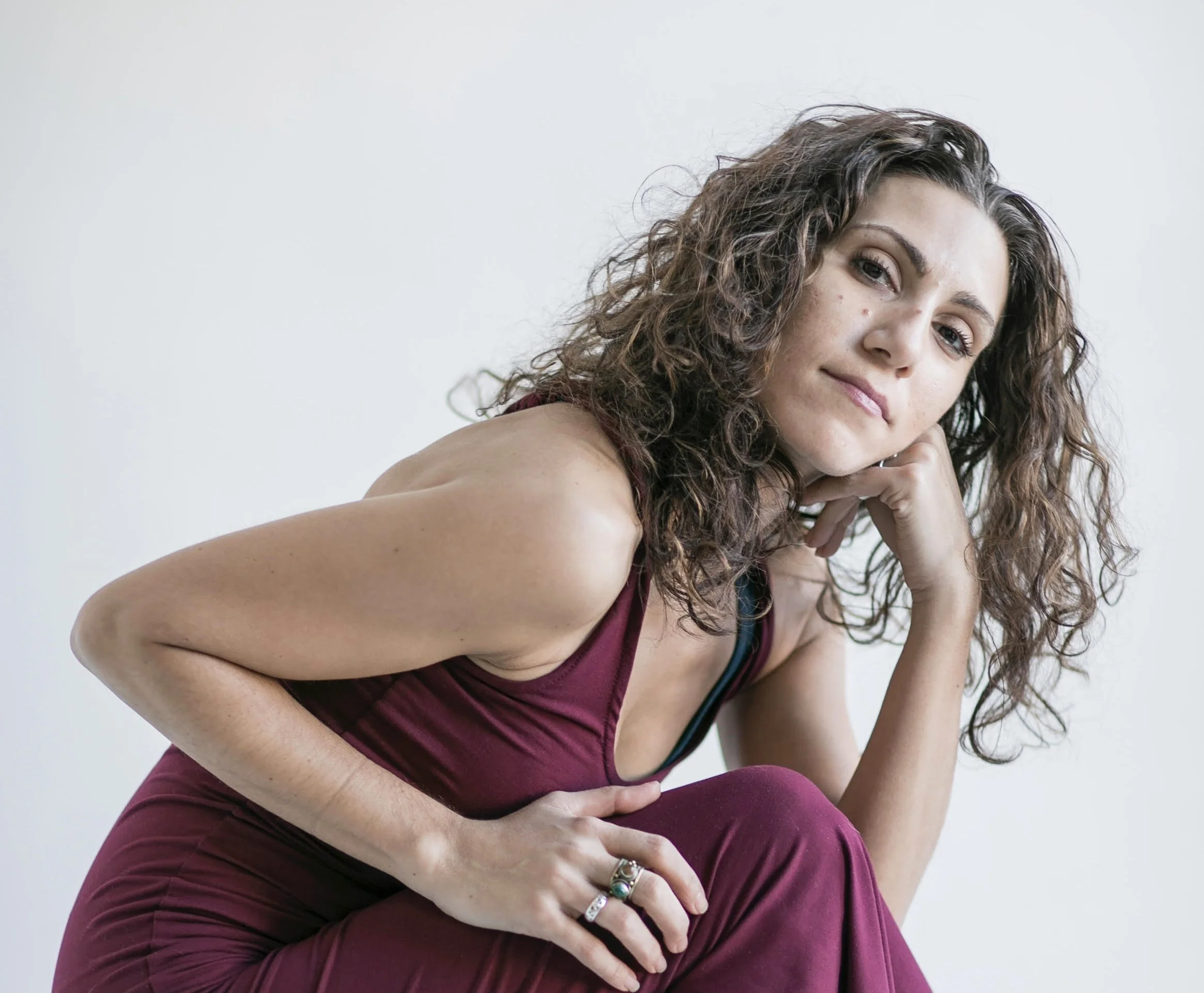 A woman with curly brown hair and light skin, sitting against a plain white background, wearing a sleeveless maroon top and pants, resting her chin on her hand and looking at the camera.