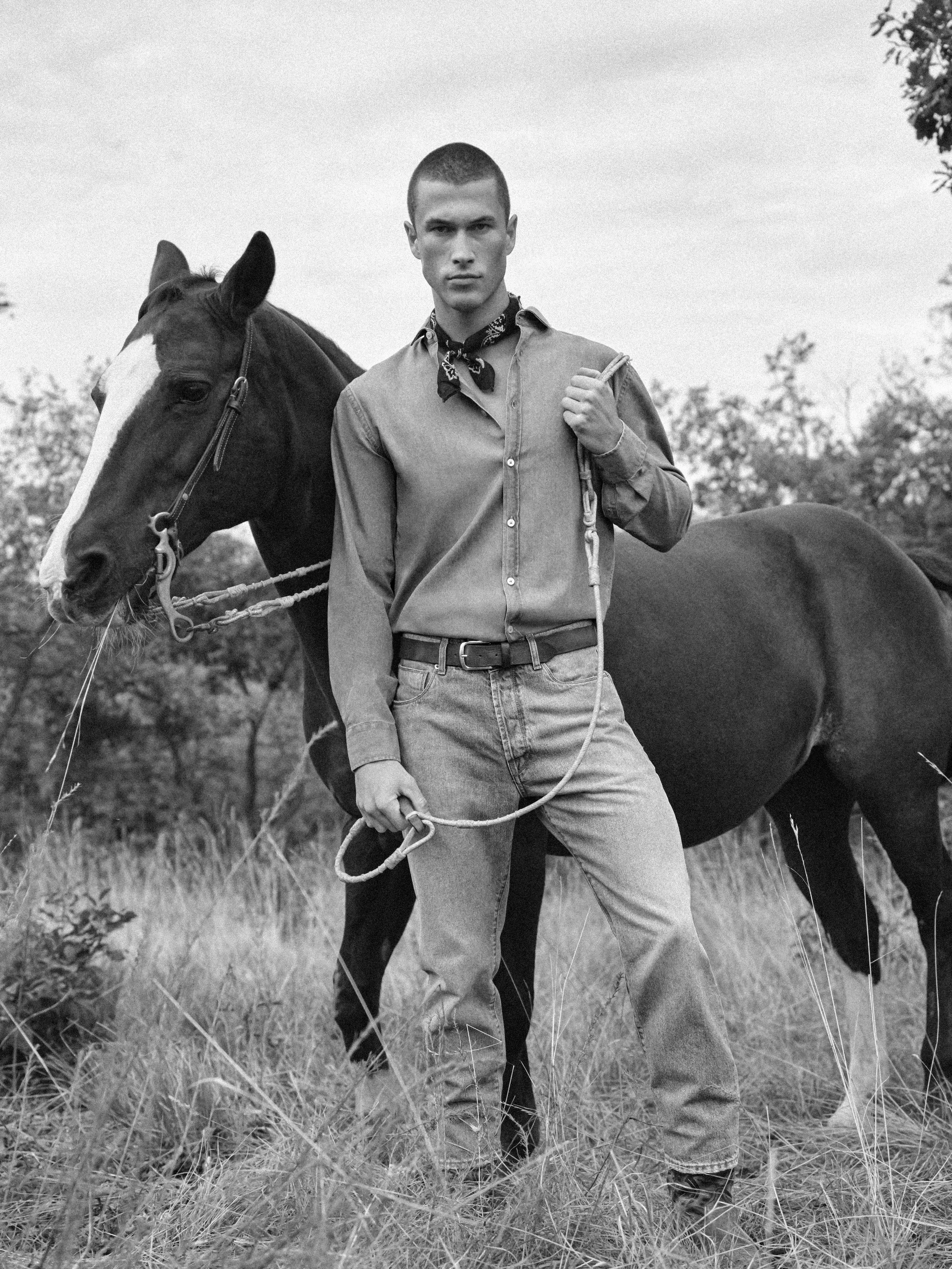 Black and white photo of a young man standing in a field with tall grass, holding a rope attached to a horse. The young man is wearing jeans, a button-up shirt, and a bandana around his neck. The horse has a dark coat with a white stripe on its face.