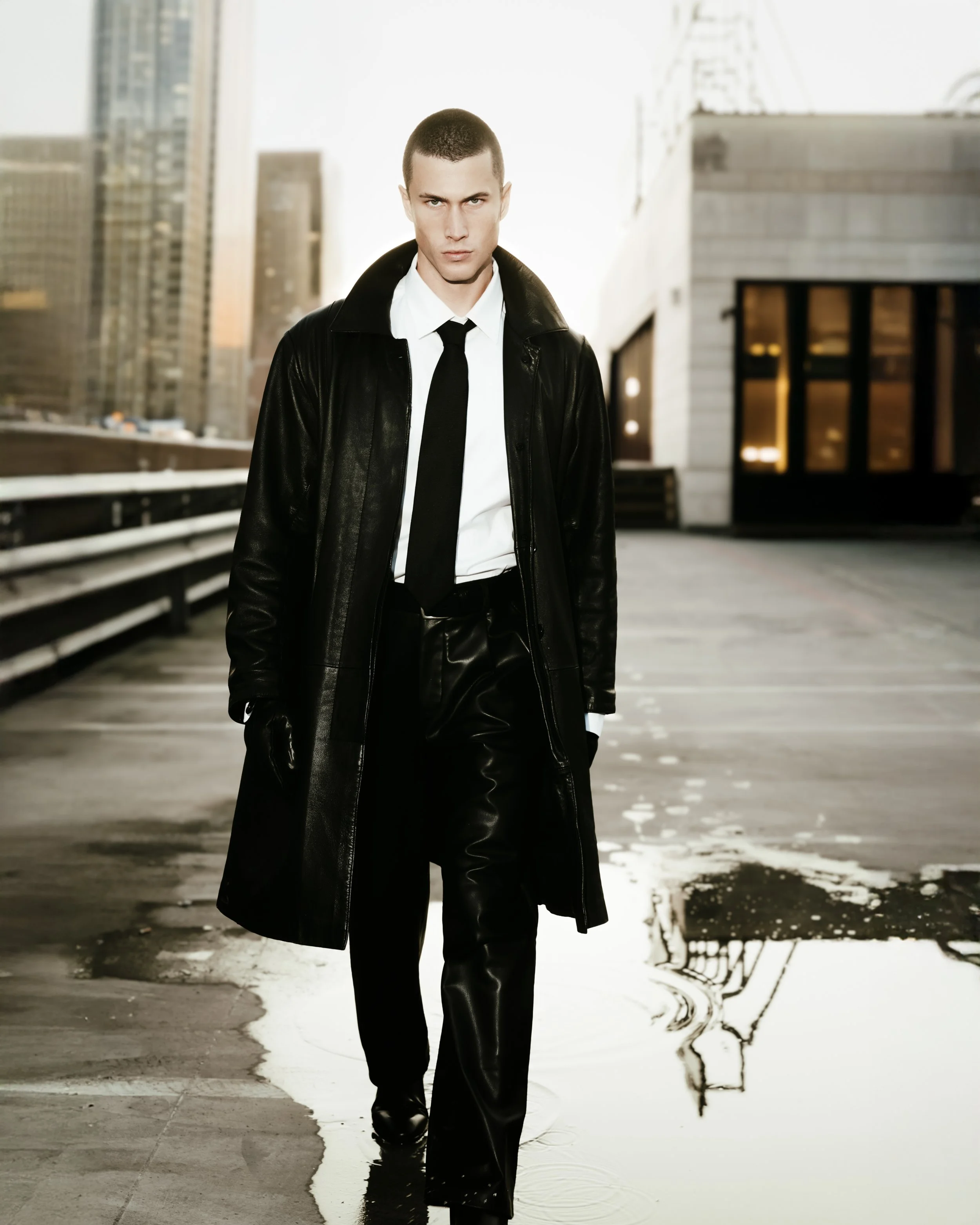A young man with short hair and a serious expression, wearing a black leather trench coat, white shirt, and black tie, walking on a rooftop or parking lot with city buildings in the background during sunset or sunrise.