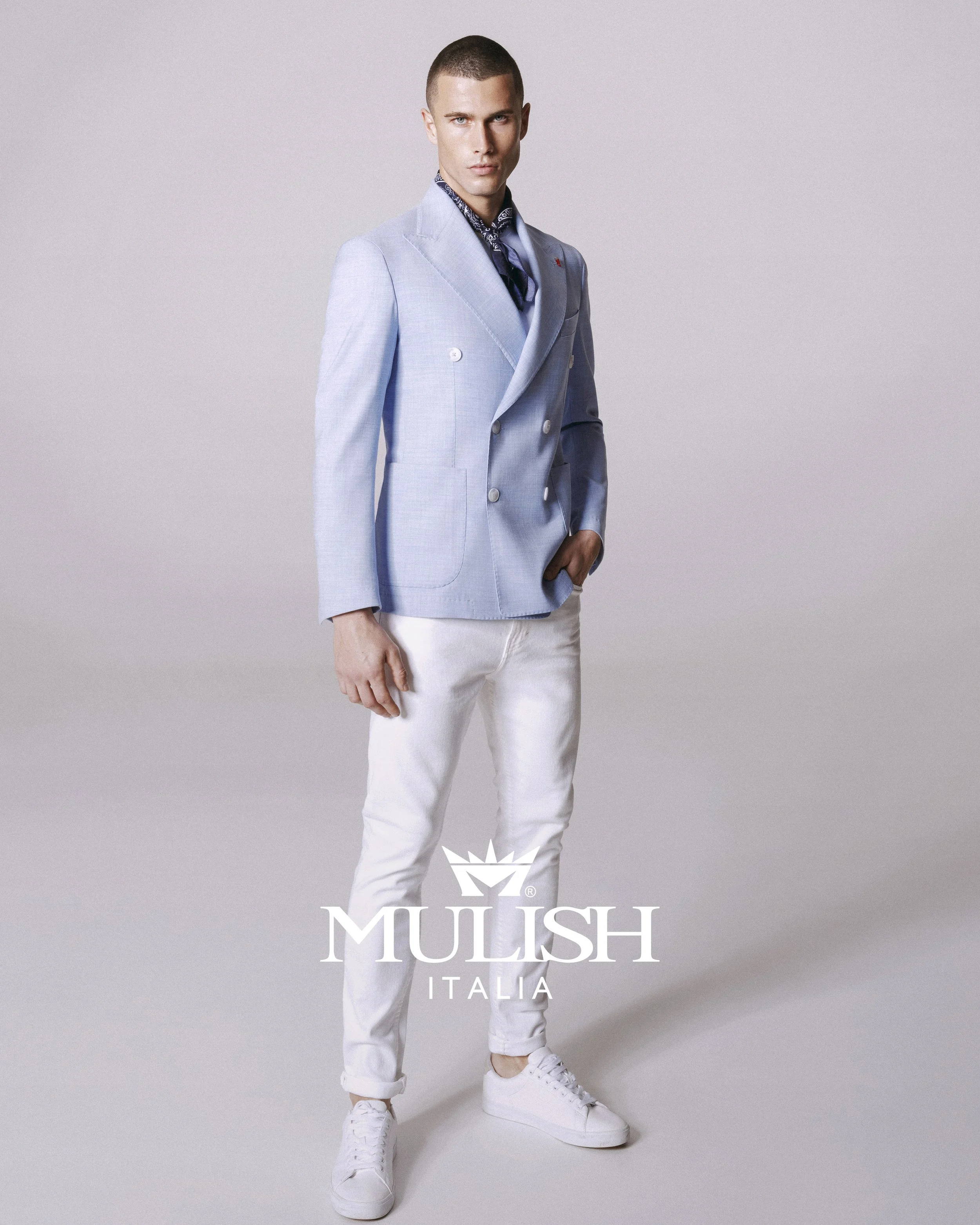 A man wearing a light blue double-breasted blazer, a dark patterned bandana around the neck, light-colored pants, and white sneakers, standing against a plain background with the words "MUSH ITALIA" and a crown logo at the bottom.