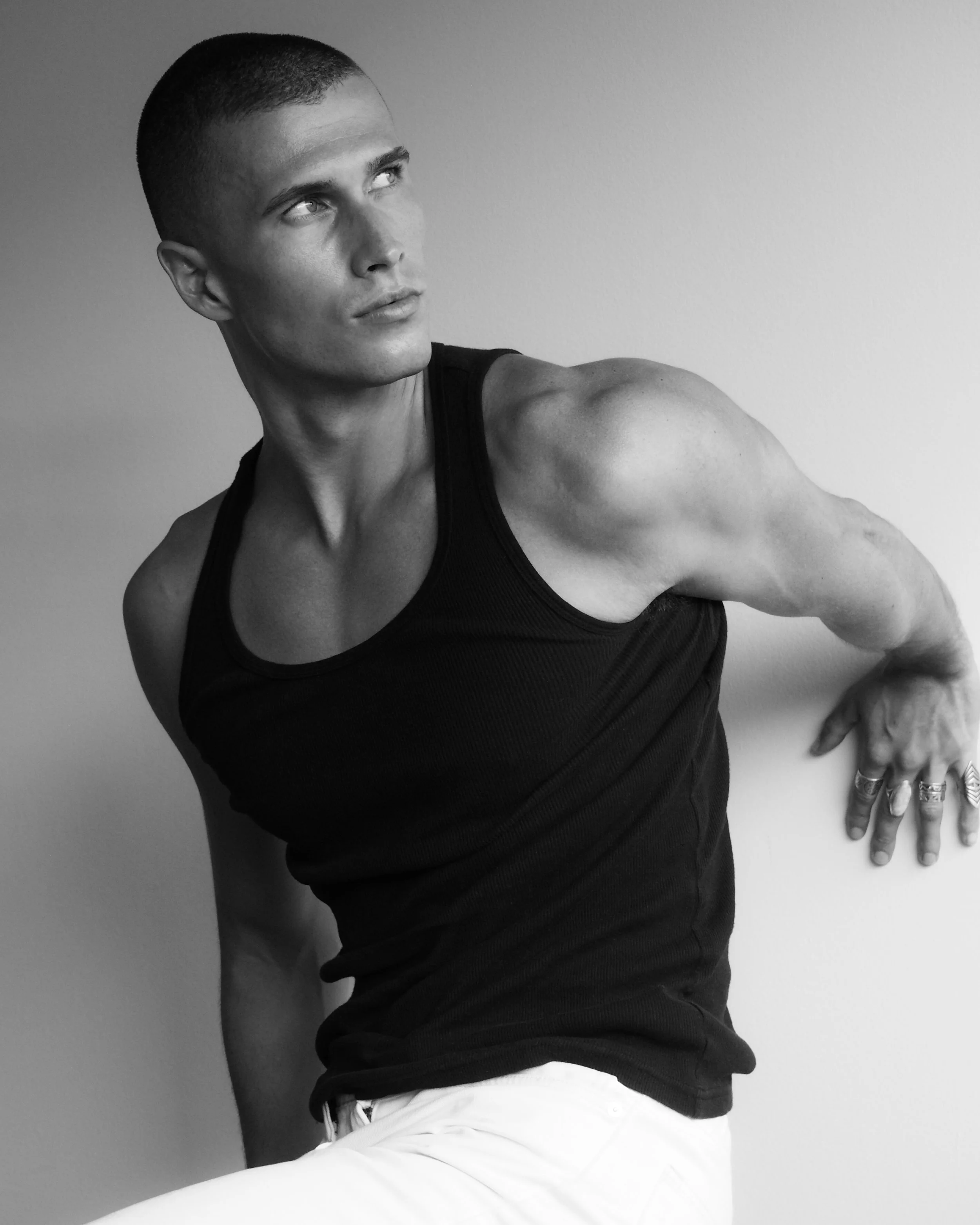 Black and white photo of a young man with a buzz cut hairstyle, wearing a sleeveless black shirt and white shorts, positioned against a plain wall, looking off to the side.