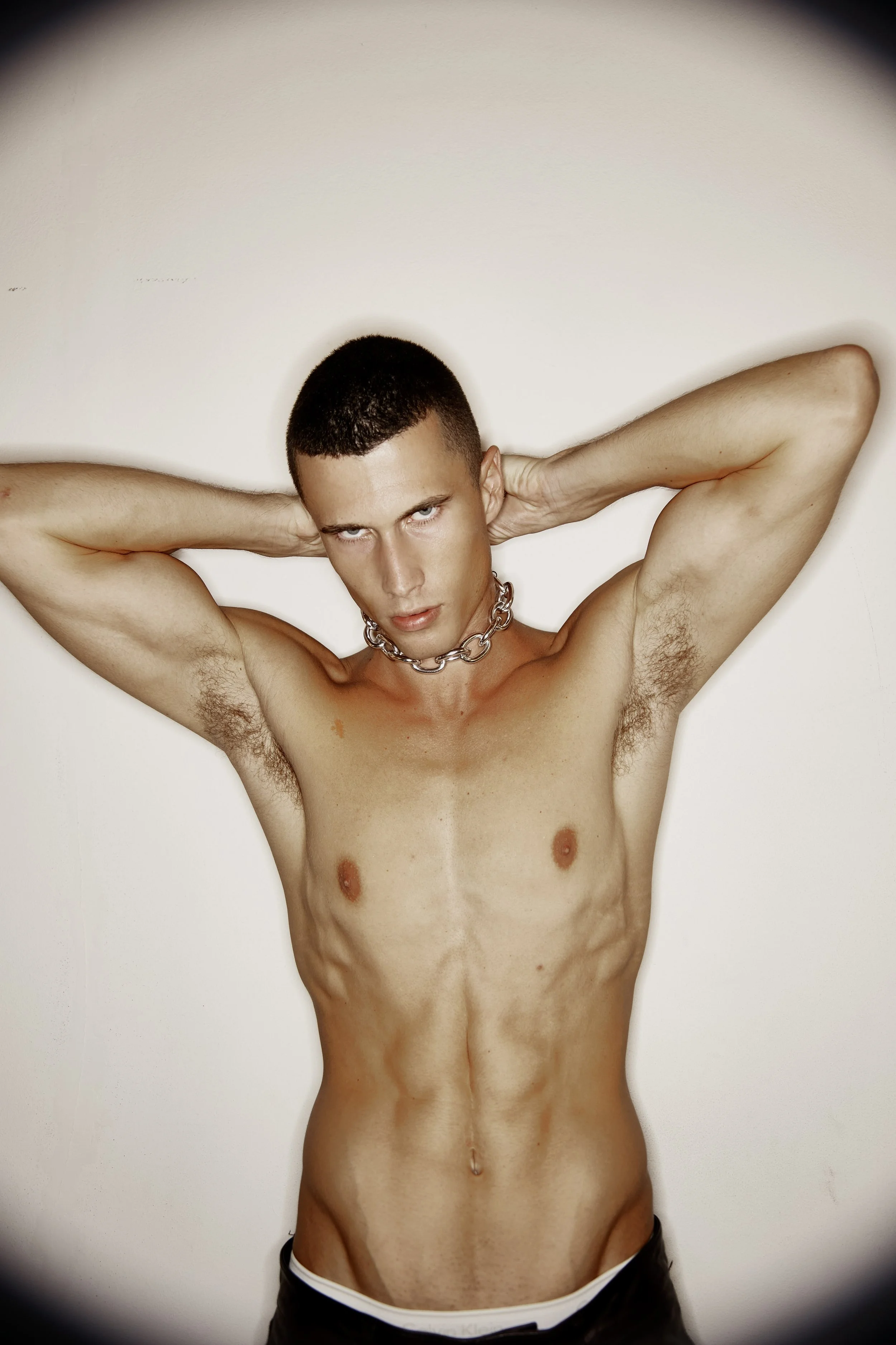 A shirtless man with short dark hair and light skin, wearing a thick silver chain necklace, posing against a plain white background. He has his arms raised behind his head and is looking directly at the camera with an intense expression.