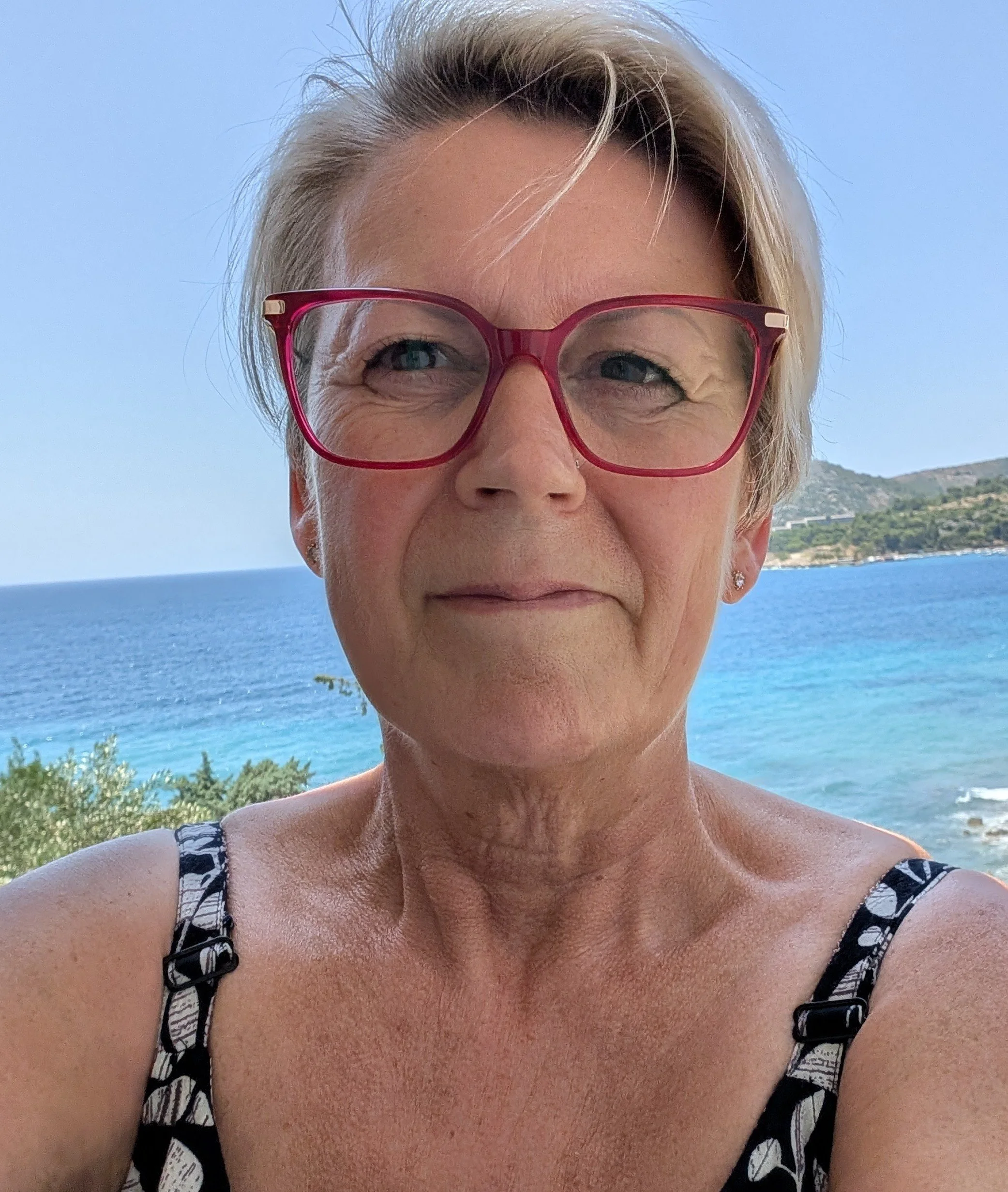 Karen Woodall smiling, who has short blonde hair, is wearing glasses, a patterned black top, and is looking directly at the camera against a bright blue sky and ocean background.