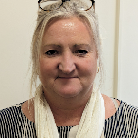 Biddy Mayo smiling, who has short blonde hair and is wearing a white scarf and a black stripey top, looking directly at the camera against a white background.