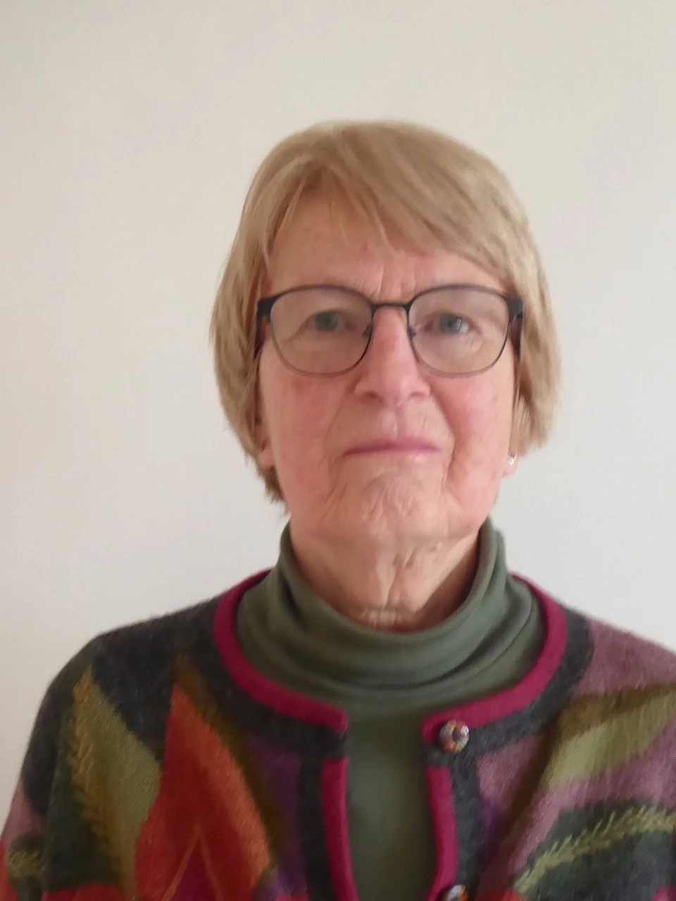 Gillian Pawson smiling, who has short blonde hair, is wearing glasses, a green jumper, a black patterned jacket and is looking directly at the camera against a white background.
