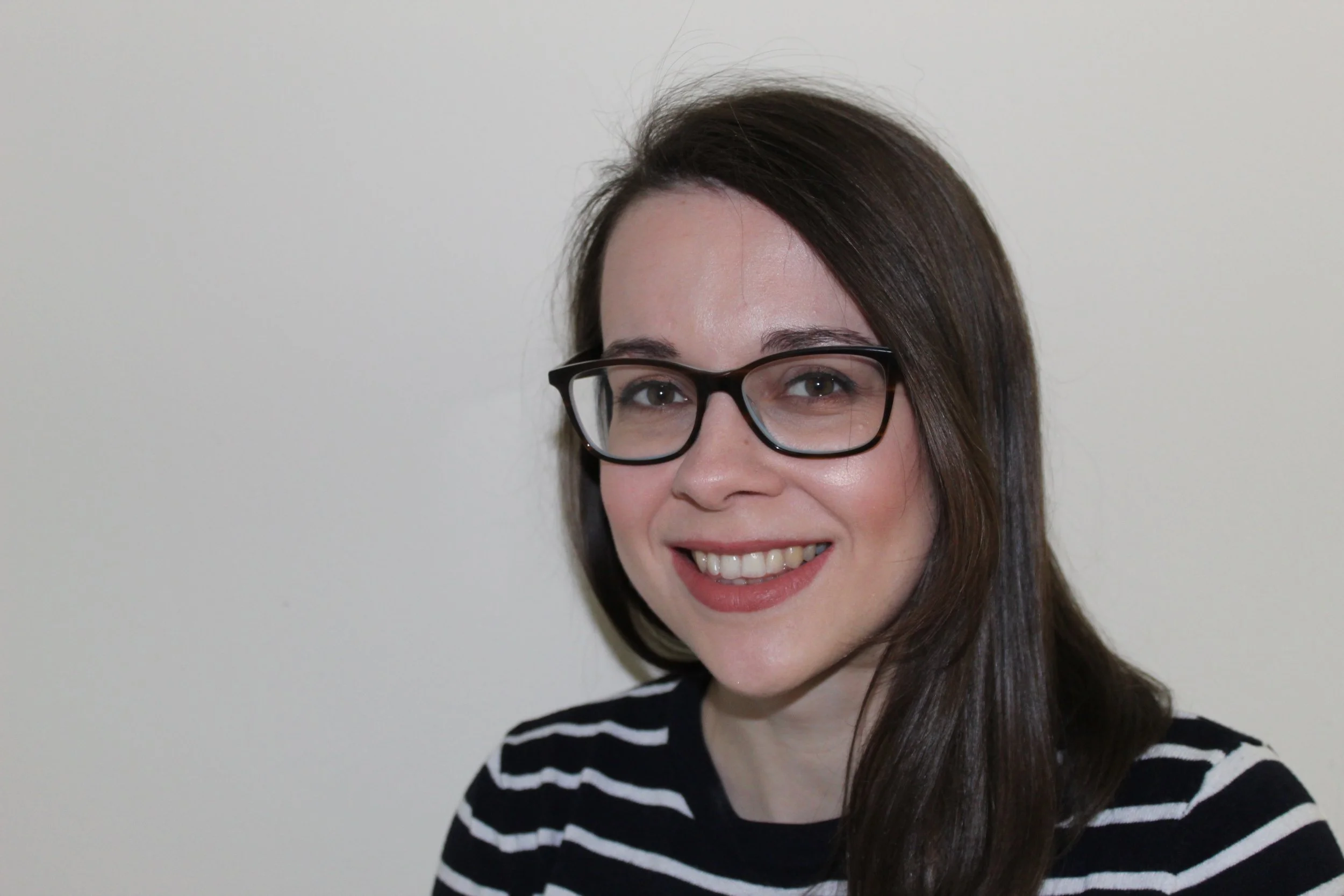 Headshot of Katherine Cronnelly who has shoulder length brown hair over one shoulder, is wearing glasses and a stripey black and white top..