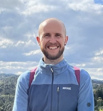 Callum Dixon smiling, who is bald with a beard and wearing a blue jacket looking directly at the camera with a bright sky in the background.