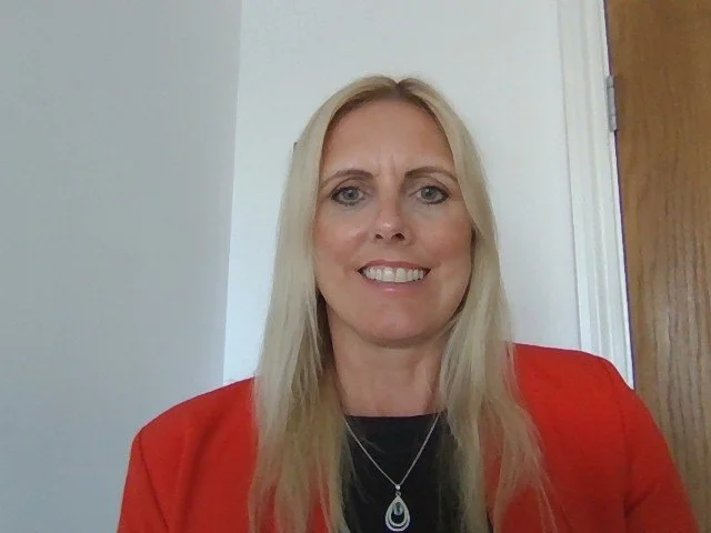 Marcella Booth smiling, who has long blonde hair and is wearing a red blazer and black top, looking directly at the camera against a white background.