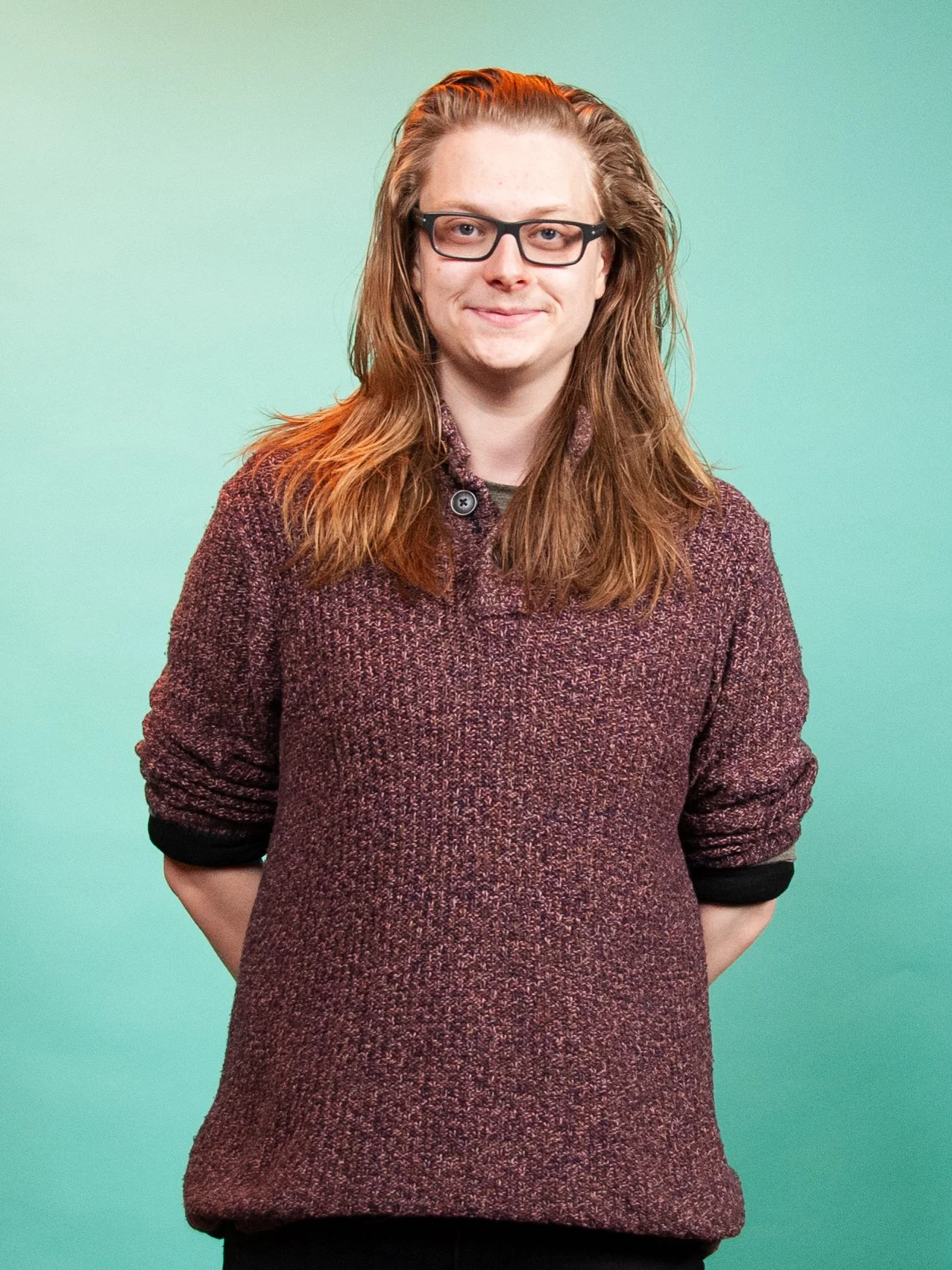 Matthew Henderson smiling, who has shoulder length brown hair, is wearing glasses, a burgandy jumper, and is looking directly at the camera against a green background.