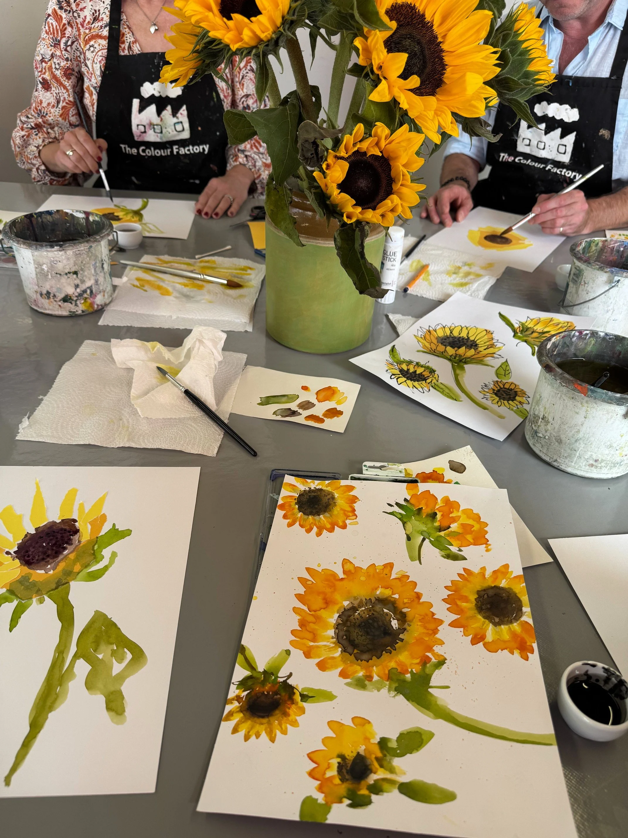 Students painting loose watercolour and ink sunflowers at a workshop table at The Colour Factory, surrounded by fresh sunflowers and painting materials.