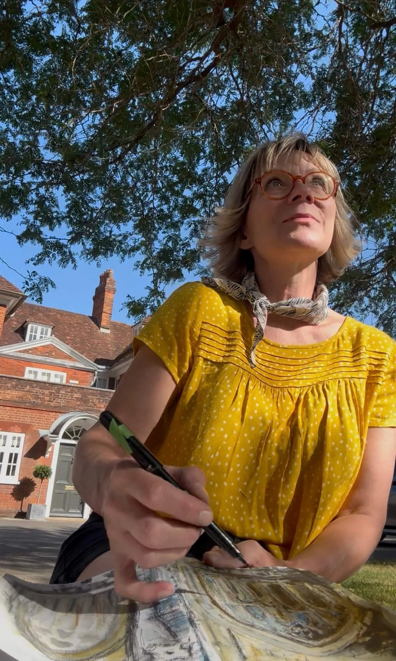 A woman sits under a tree sketchin by an Georgian house in Winchester