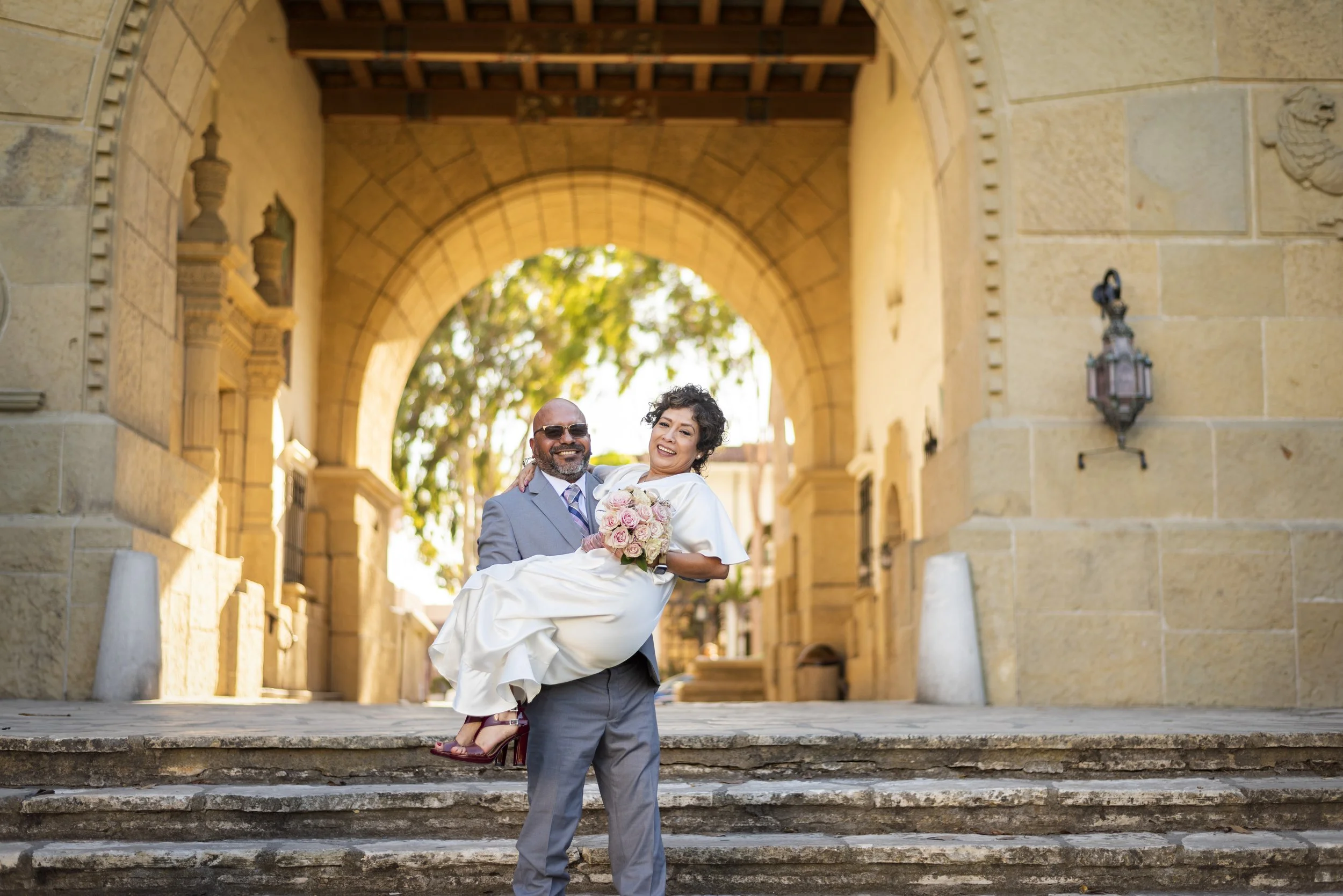 A man in a gray suit holding a smiling woman in a white dress and holding a bouquet of pink roses, in front of an arched stone building.