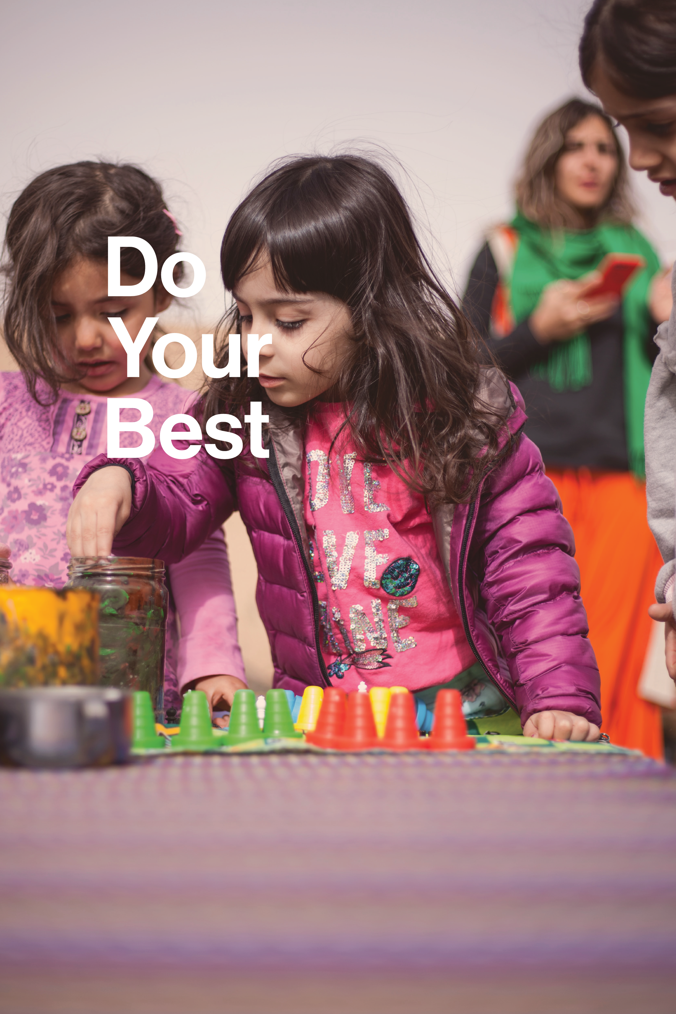 Children playing a game with colored cups on a table, with a woman using her phone in the background.