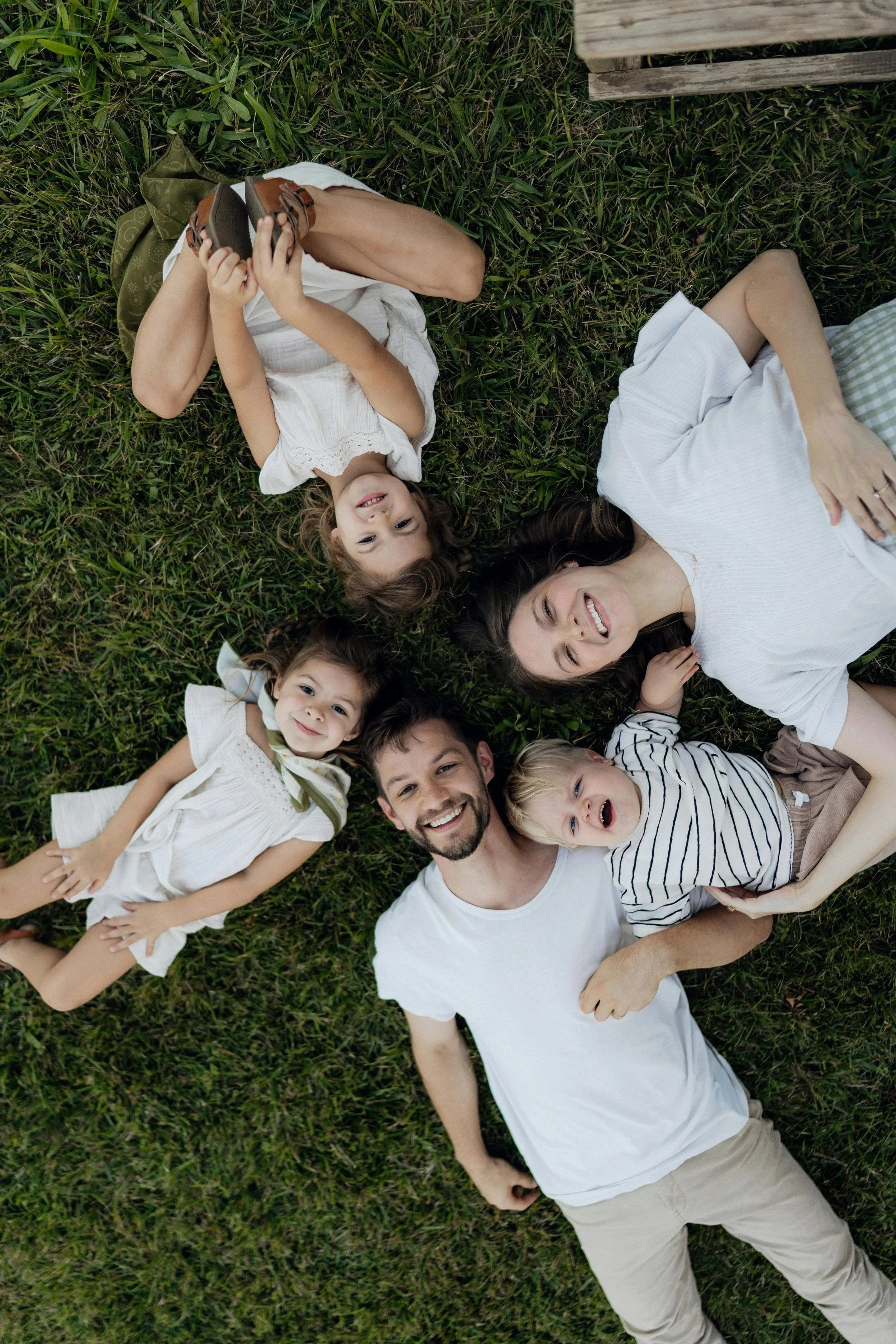 A family of six lying on green grass, smiling and looking up at the camera, with some children holding shoes.