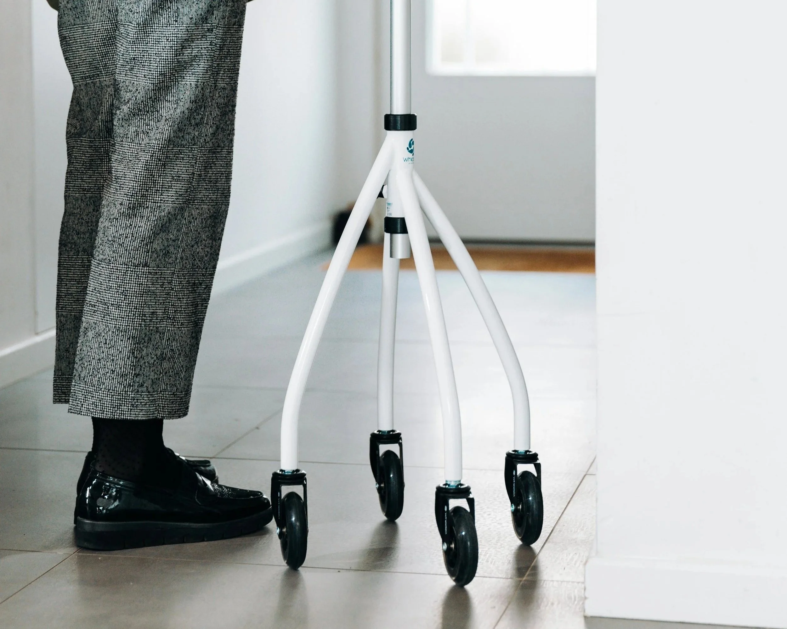 Person wearing black dress shoes and patterned pants standing next to a white walker with four wheels, inside a room with tiled floor and white walls.