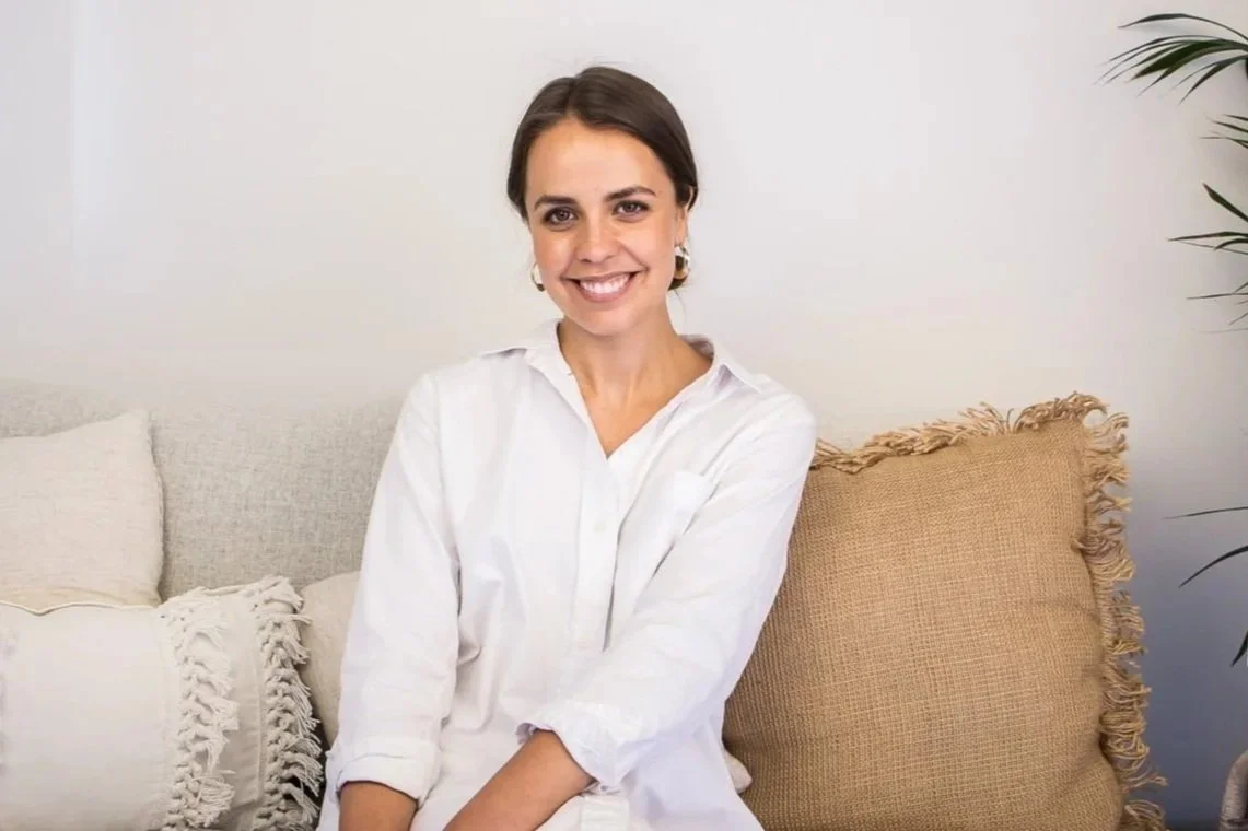 A woman with brown hair, wearing a white shirt, sitting on a sofa with beige and white throw pillows, smiling in a bright living room with a plant in the background.