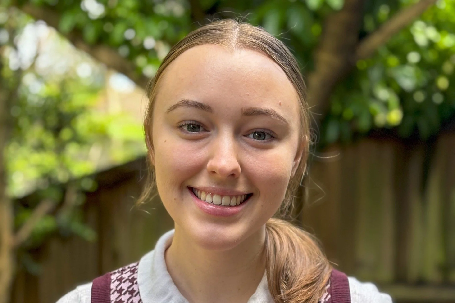 A young woman smiling outdoors with greenery and a wooden fence in the background.