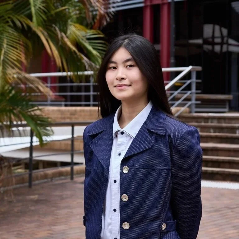 A young woman with long black hair wearing a navy blazer and white button-up shirt, standing outdoors in front of a railing and tropical plants.