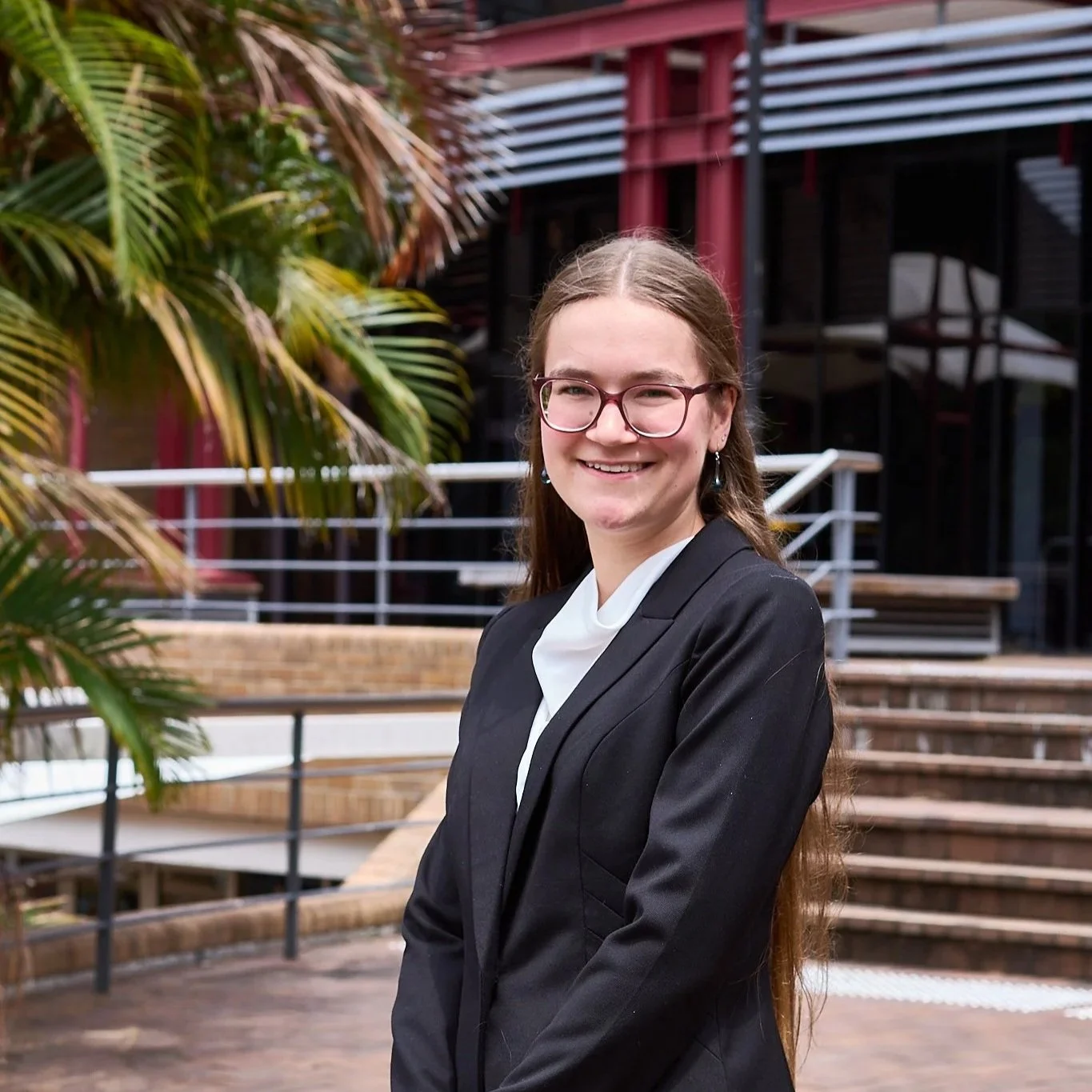 A young woman with glasses and long hair, dressed in a black blazer and white blouse, smiling outdoors with plants and modern building architecture in the background.