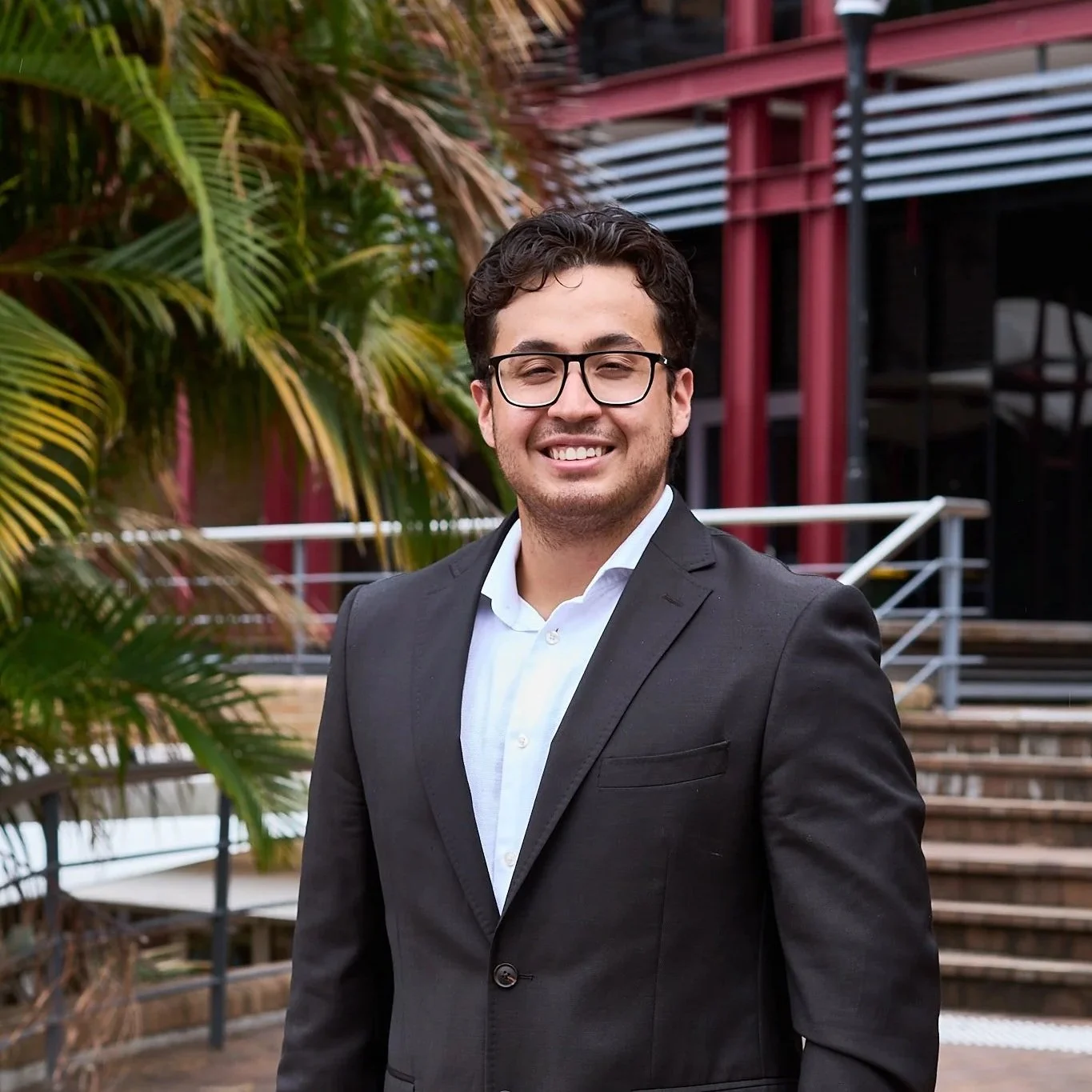 A young man with dark hair, wearing glasses and a black suit, smiling and standing outdoors near steps and palm trees.