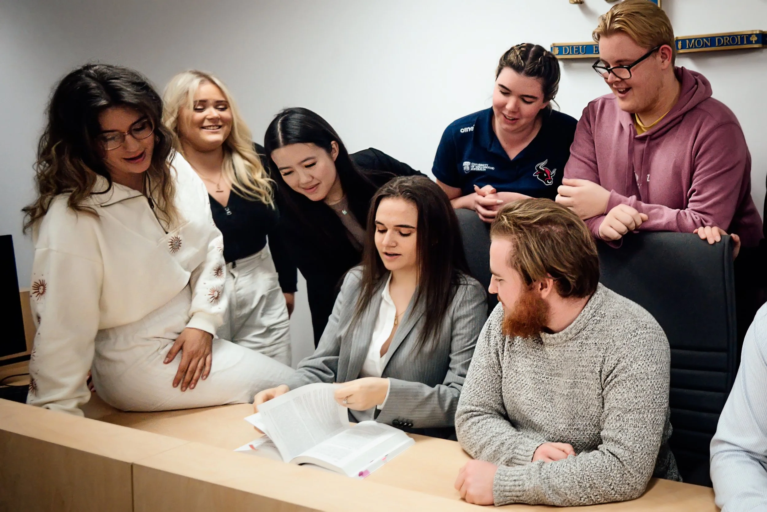 A woman with long dark hair in a gray blazer and a man with a beard and gray sweater are sitting at a desk looking at a document, surrounded by five women standing close, observing and smiling, in a room with white walls and a religious banner.