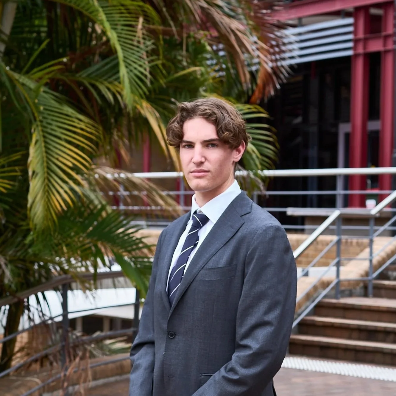 A young man in a gray business suit and striped tie standing outdoors near tropical plants with modern red metal structures in the background.