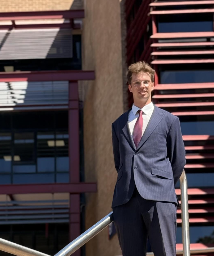 A man wearing a blue suit with a pink tie standing outdoors in front of a building with red architectural details and windows.