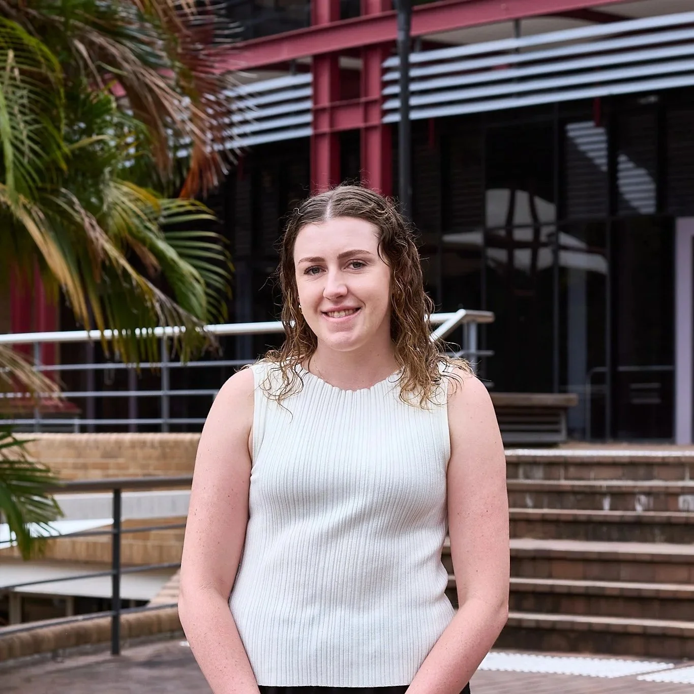 A woman with curly brown hair wearing a white sleeveless top standing outdoors near a staircase and a large green plant, with a modern building featuring glass windows and red structural elements in the background.