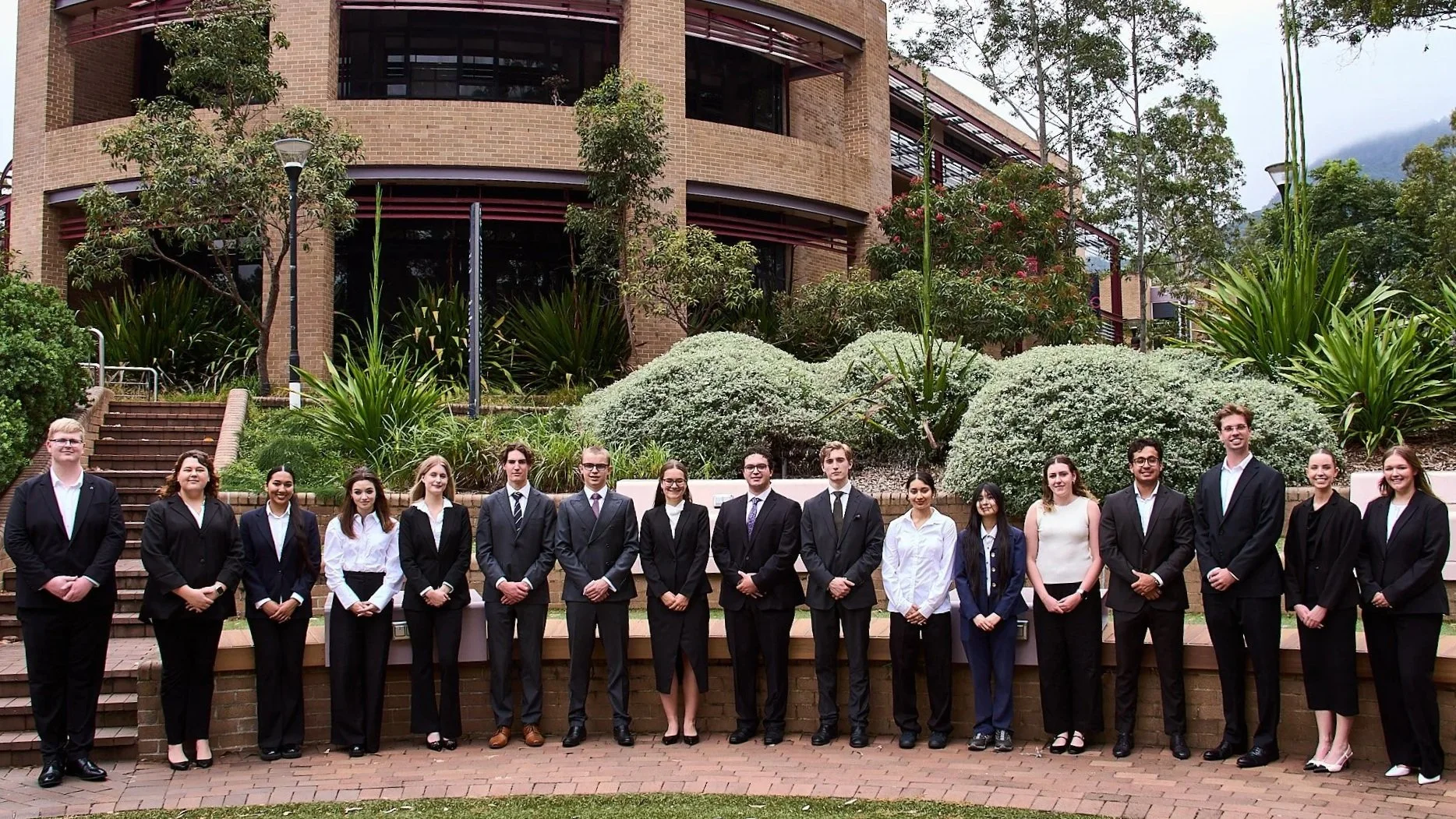 Group of 15 young adults dressed in formal business attire, standing outdoors in a line in front of landscaped bushes and trees with a brick building in the background.